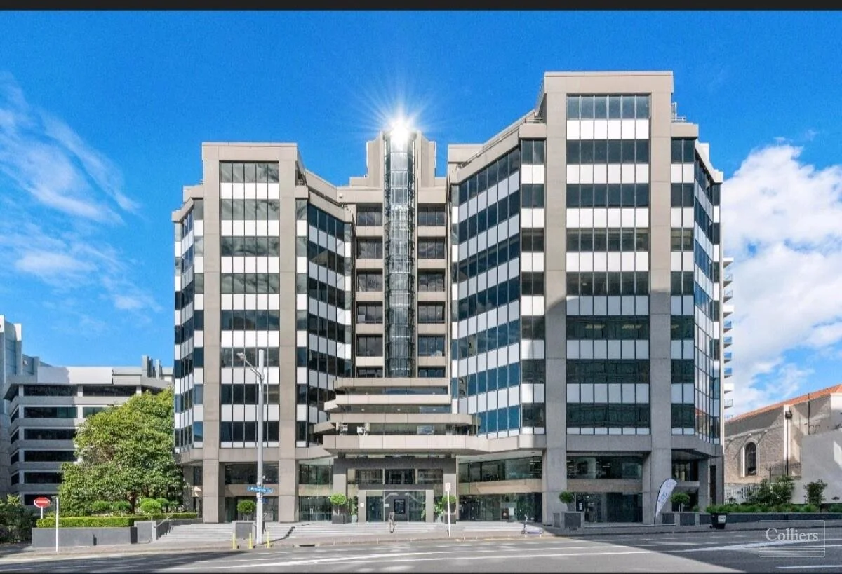 Modern multi-story office building with glass windows and a symmetrical design, located on a city street against a blue sky with some clouds.