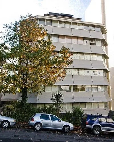 Multi-story white apartment building with curved balconies, a tree with autumn leaves in front, and parked cars at the street level.