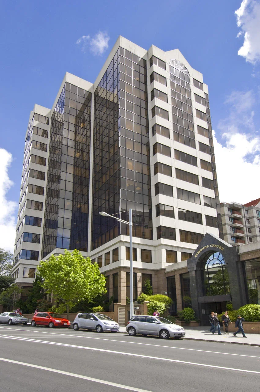 A tall modern office building with a glass facade and white concrete accents, named Diamonds Centre, with a street in front and cars parked along the curb in an urban setting under a partly cloudy sky.