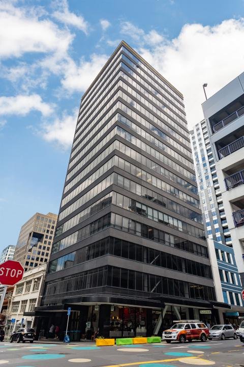 A tall modern office building with reflective glass windows on a city street, with other high-rise buildings and a blue sky with clouds in the background.