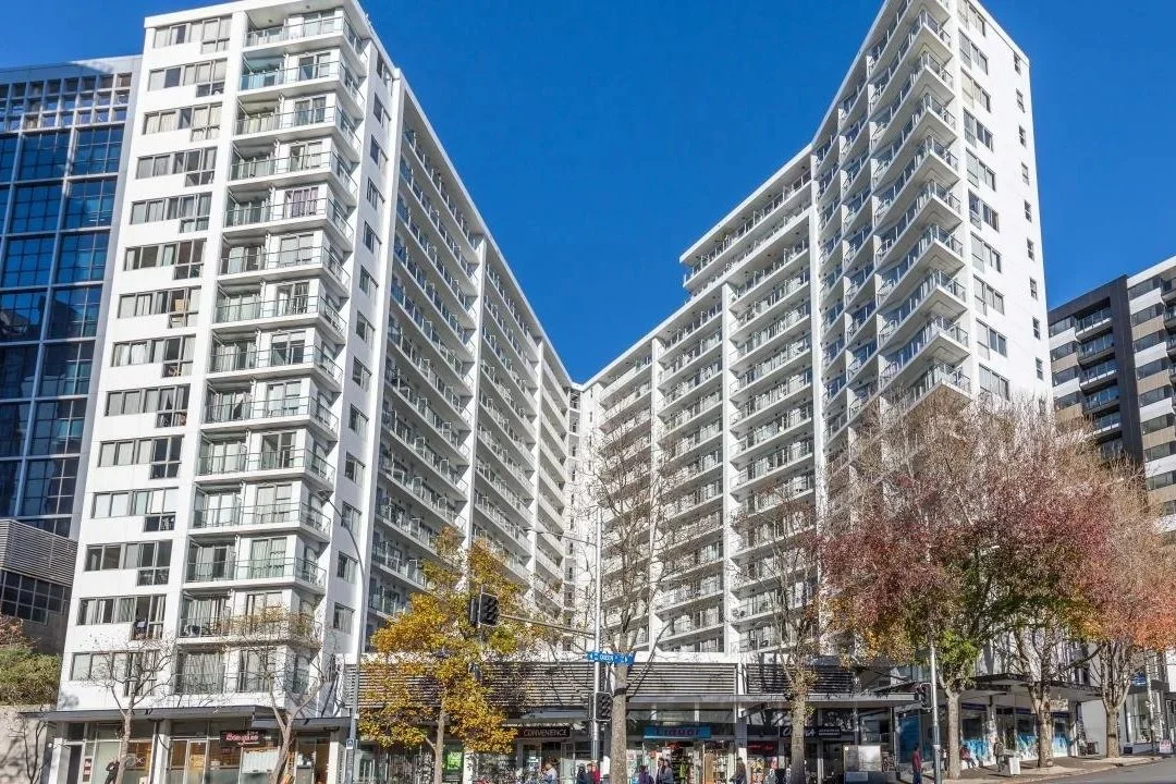 Tall white residential apartment buildings with many windows and balconies, trees with autumn foliage, and a street with pedestrians and traffic signals in the foreground.
