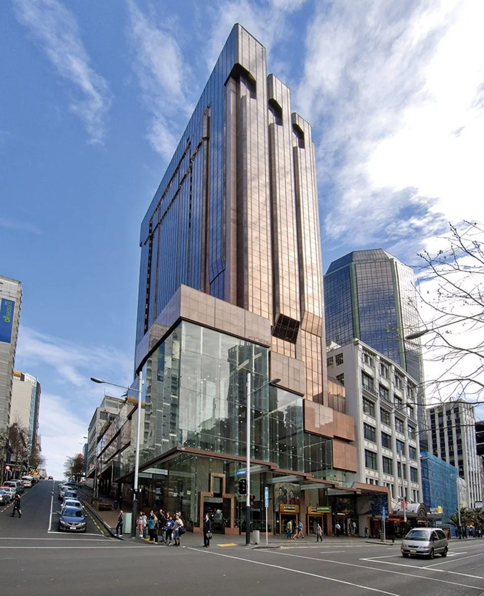 Tall modern office building with glass and copper-colored facade, located on a busy city street with cars and pedestrians, under a partly cloudy sky.
