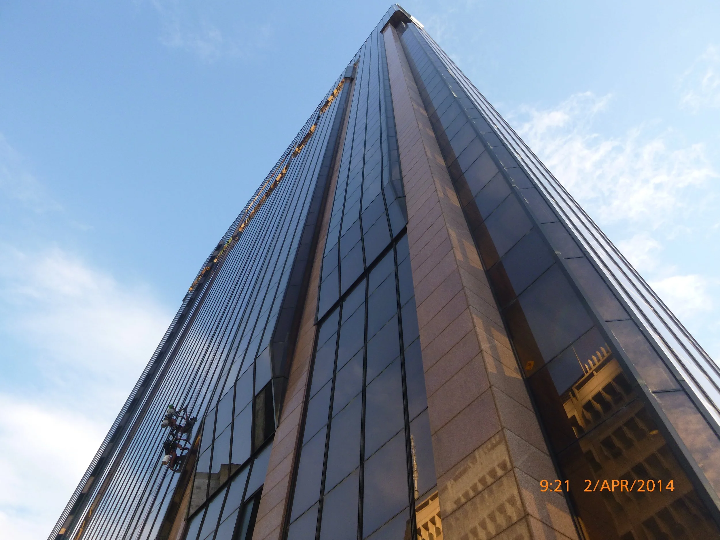 A tall glass-and-steel skyscraper taken from a low angle, reflecting the blue sky and clouds, with a timestamp of 9:21 on April 2, 2014.