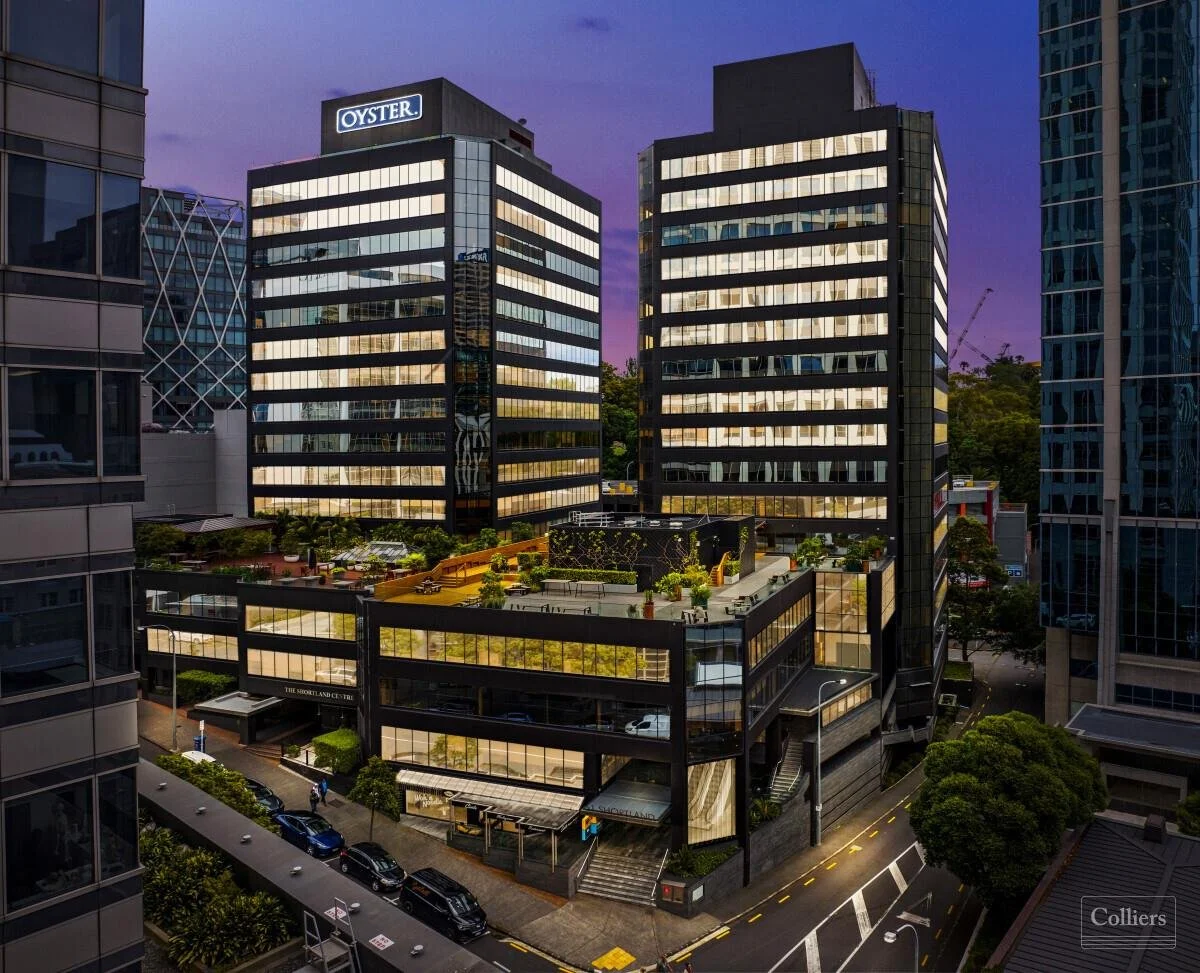 Cityscape at dusk featuring modern glass office buildings with illuminated windows, a rooftop terrace with greenery, and tree-lined streets.