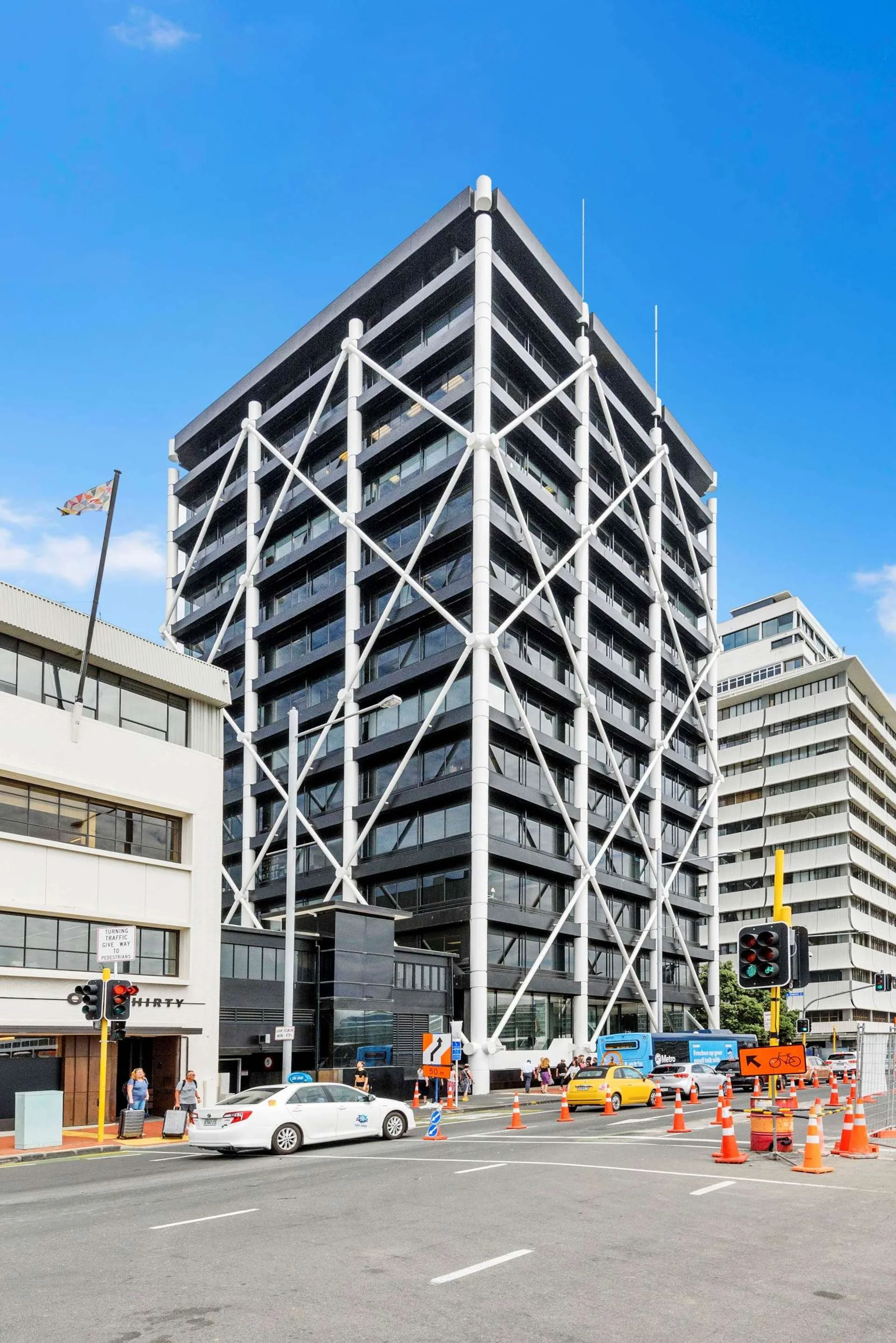 Tall modern building with a black and white exterior, supported by white diagonal braces, set against a blue sky in an urban area with traffic and pedestrians.