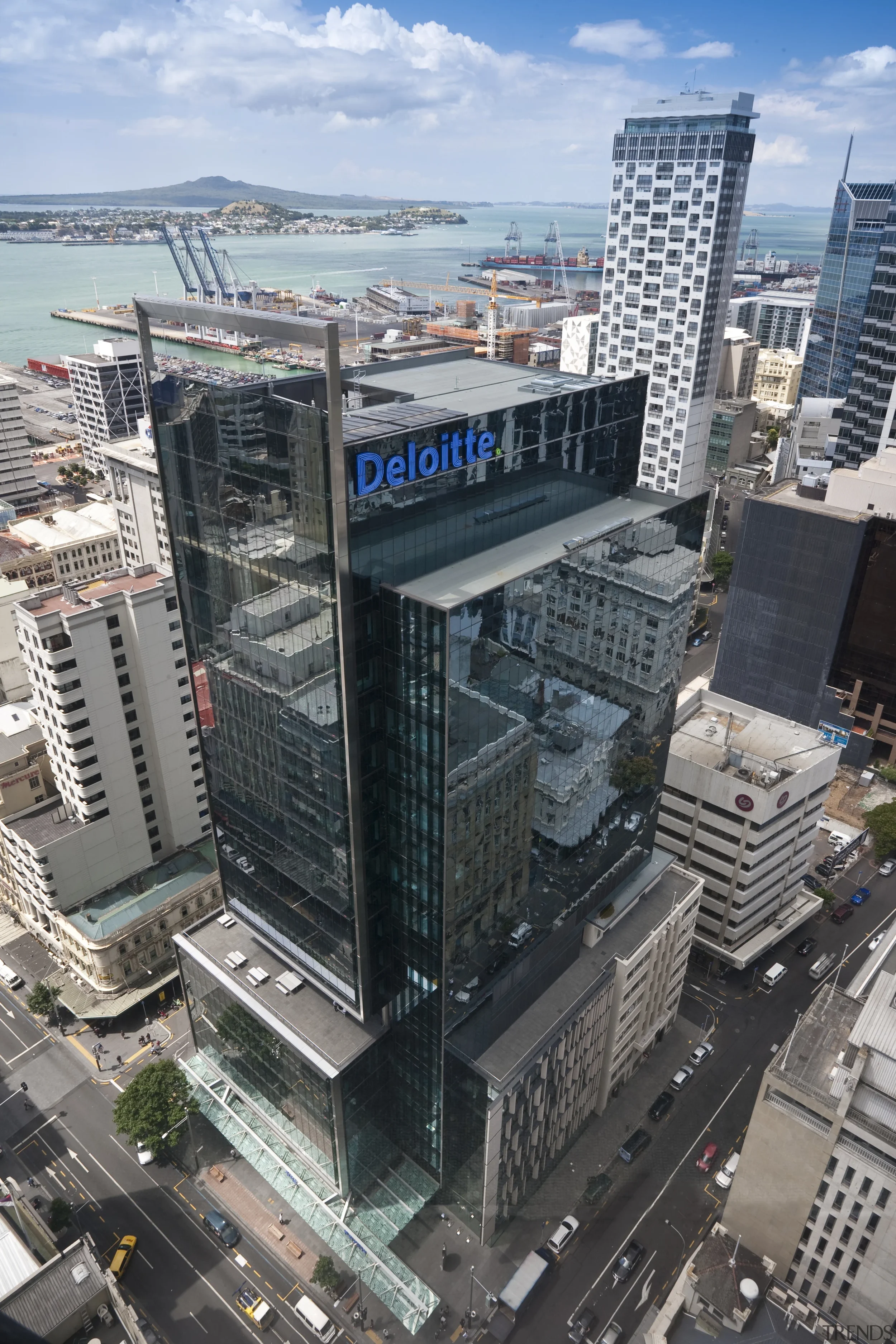 Aerial view of a modern cityscape featuring a tall glass office building with the Deloitte logo, surrounded by other high-rise buildings, streets, and a waterfront with ships and cranes in the background under a partly cloudy sky.