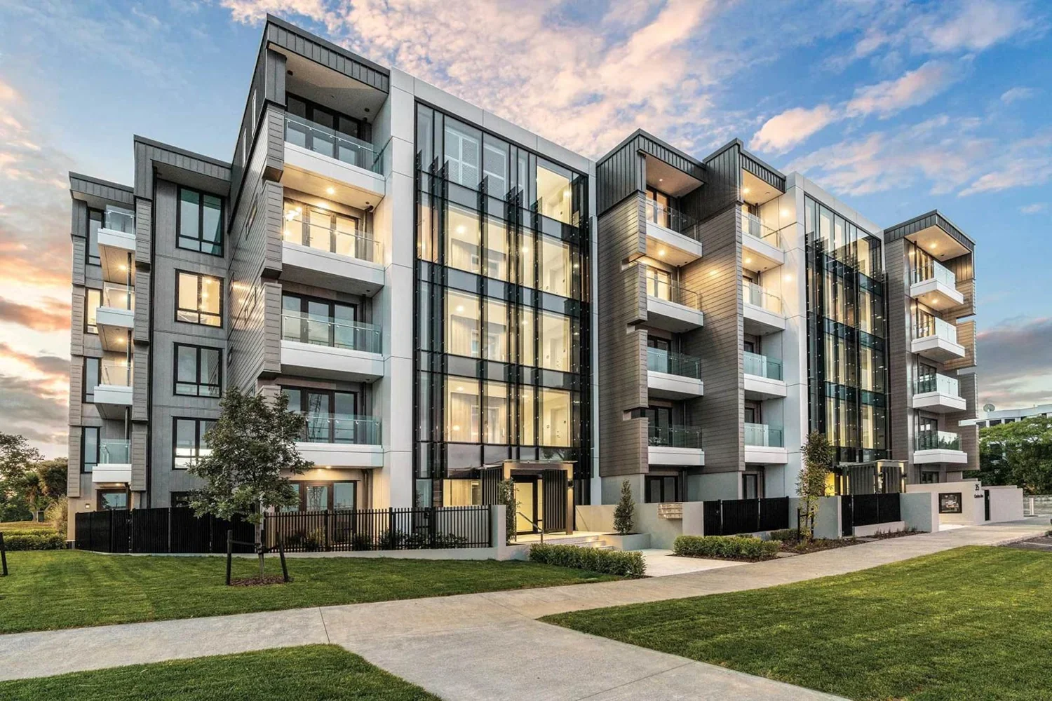 Modern multi-story apartment building with glass balconies, large windows, and landscaping in the foreground, during sunset.