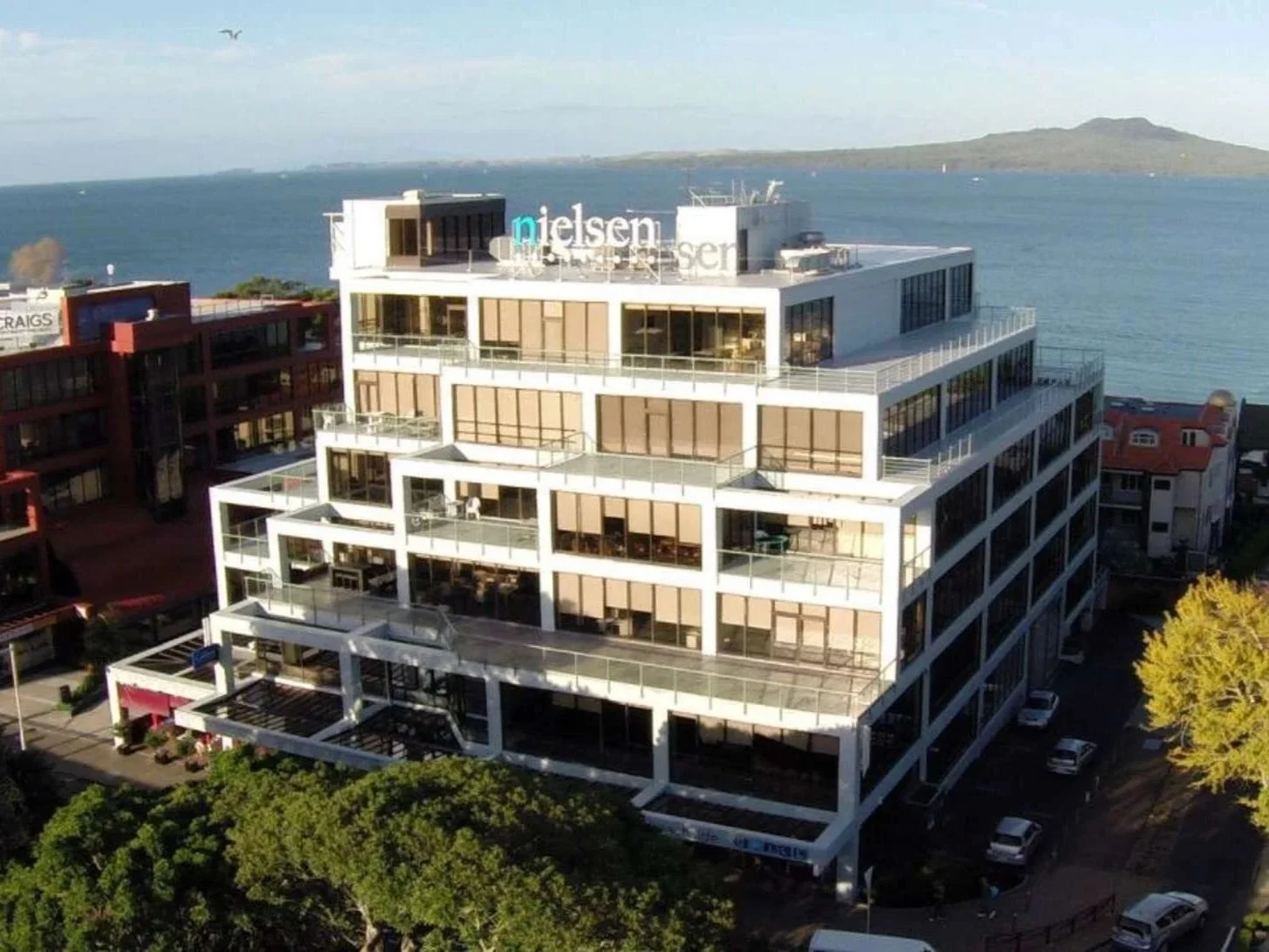 A multi-story modern white building with large glass windows, situated near the water with a distant island in the background.