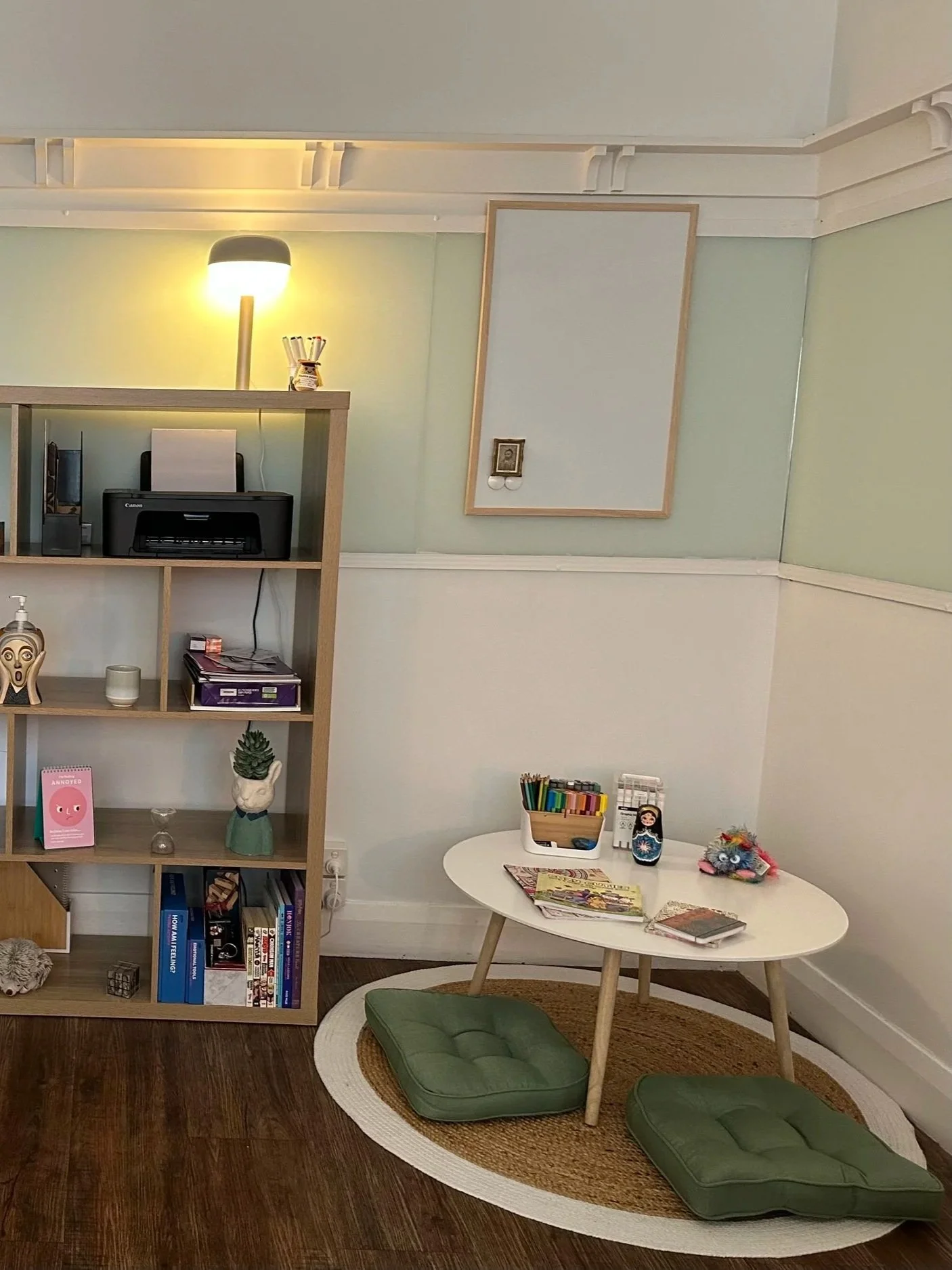 A cozy room with a wooden bookshelf, a white round table with coloring supplies and magazines, and green cushions on a circular rug on dark wood flooring. There's a small lamp, art, and decorative items.