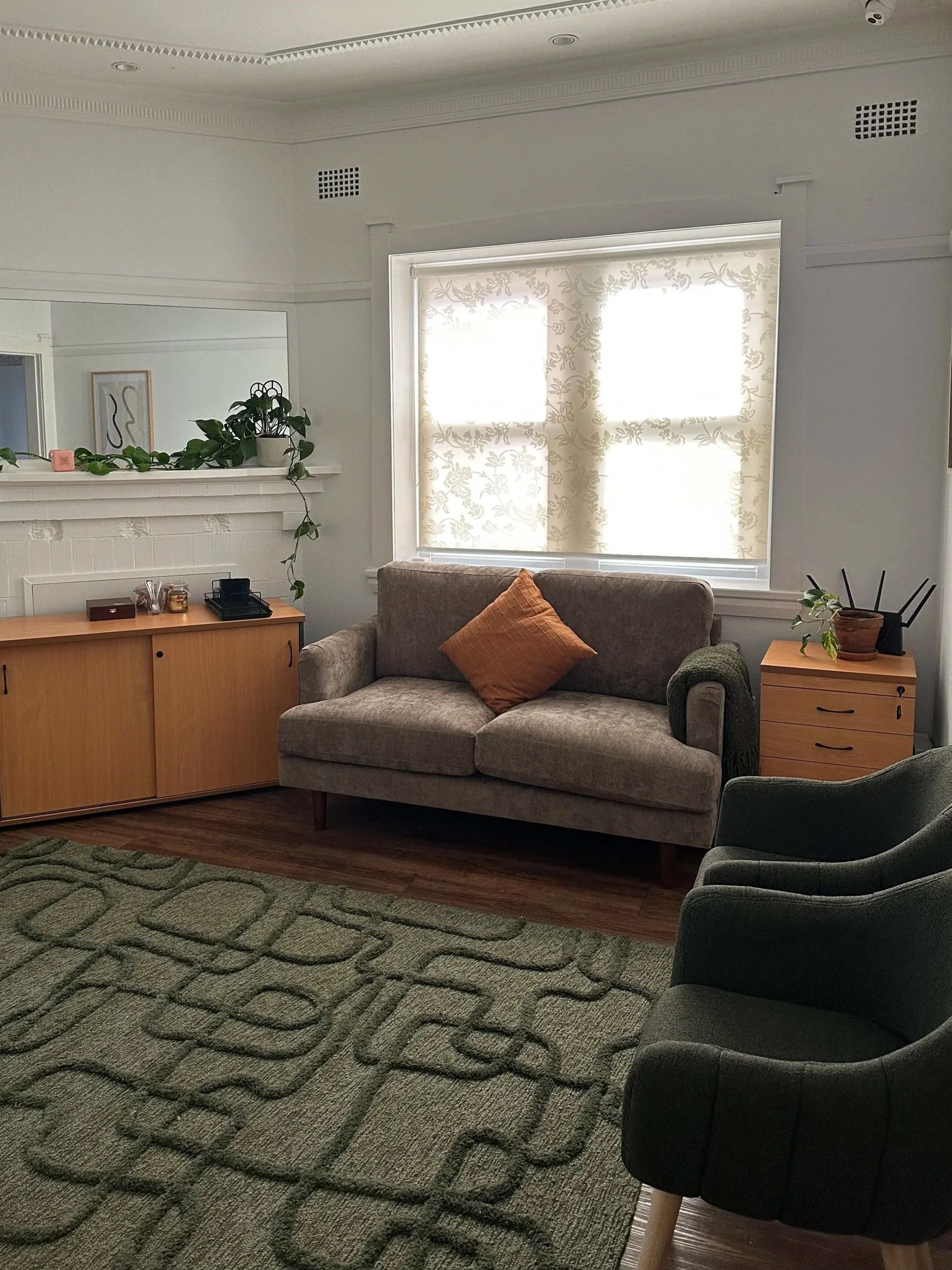 A cozy living room with a green patterned rug, a beige sofa with an orange pillow, a wooden side table with plants and decor, a fireplace with a mirror above it, a large window with patterned curtains, and a comfortable black armchair.