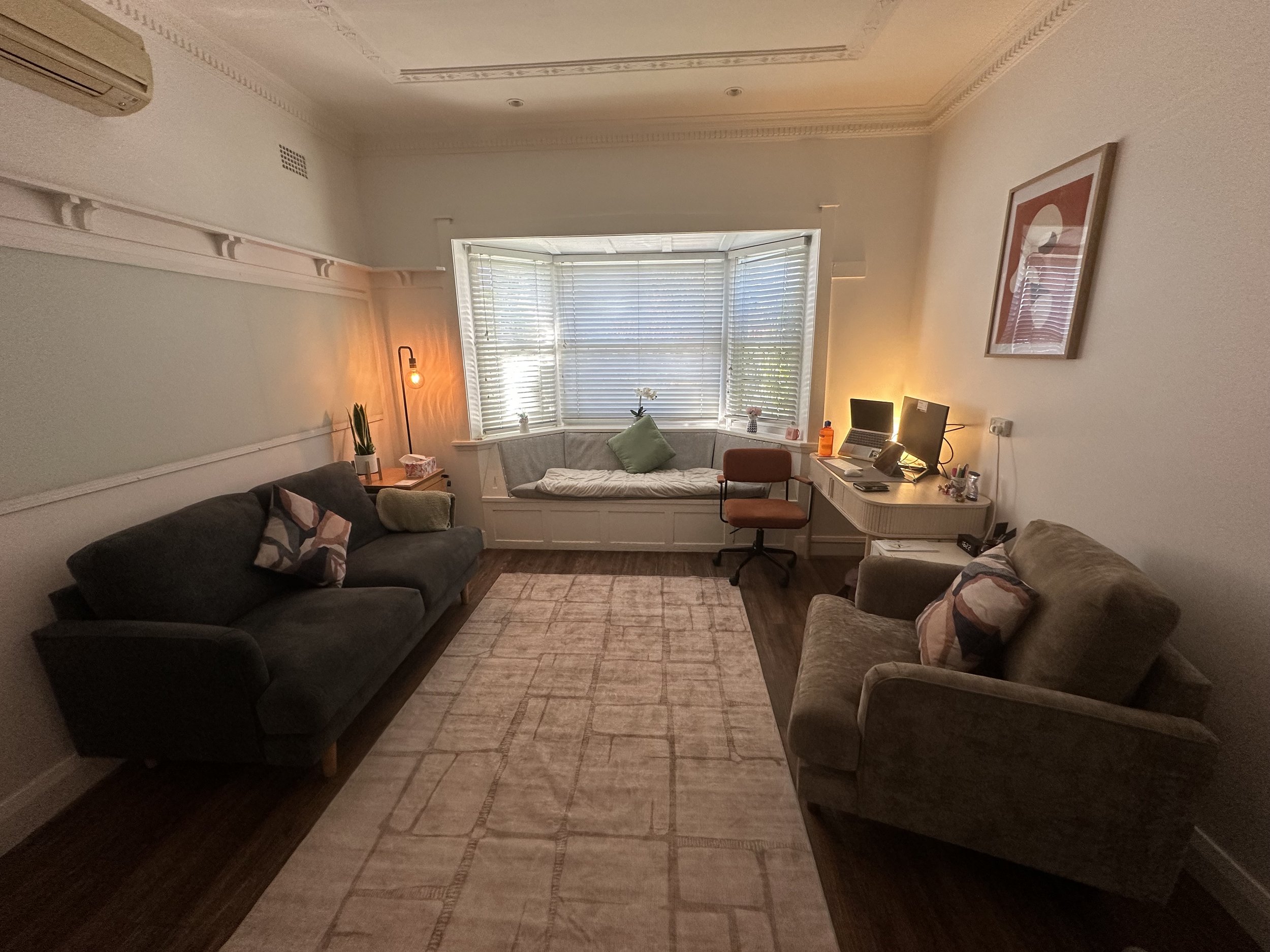 A cozy living room with a couch, armchair, and window seat with cushions, a desk with a computer, and warm lighting.
