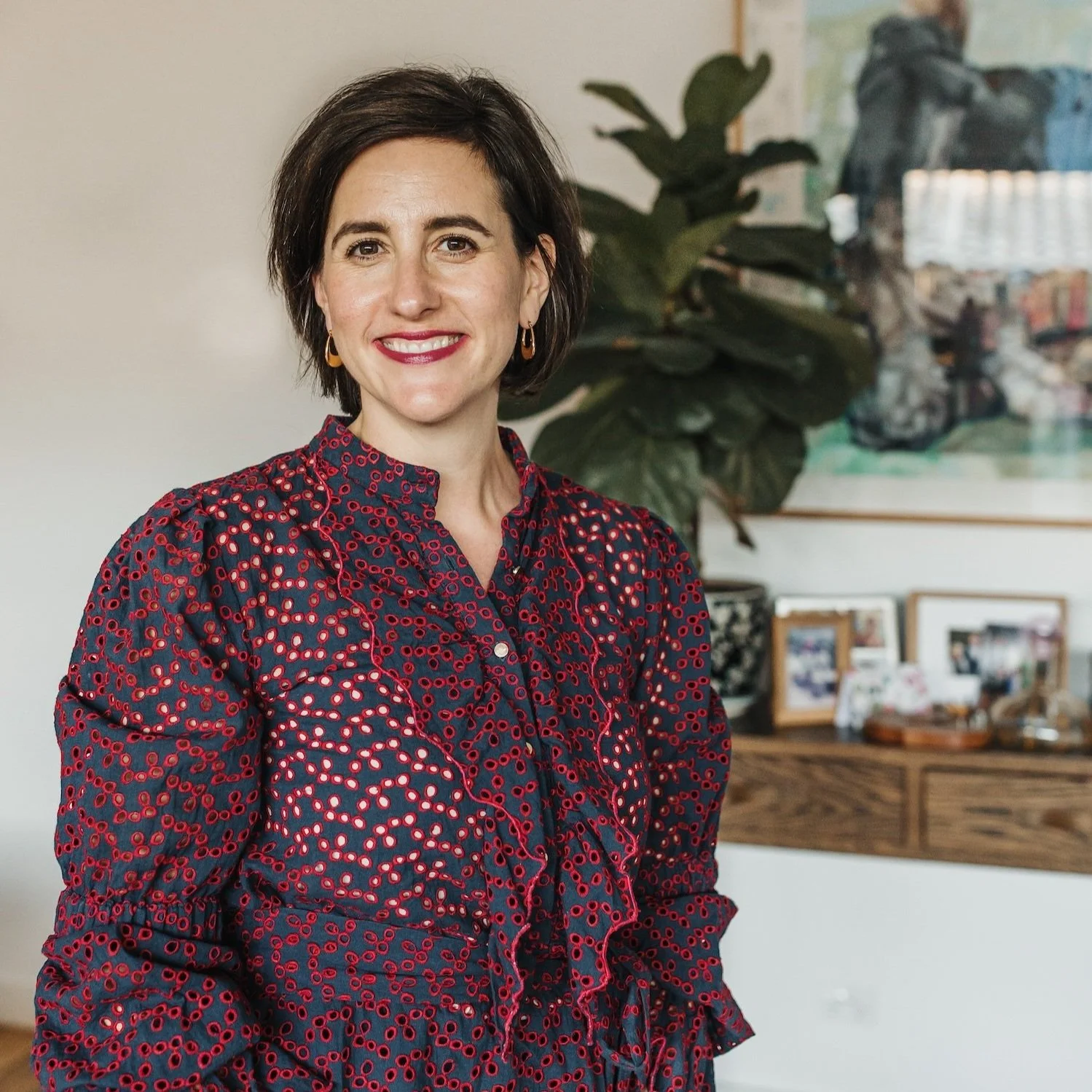 Meet Emily Lines, Aged Care Advocate. A woman with short dark hair wearing a red and navy patterned blouse, standing indoors with a large houseplant and a wall art in the background.