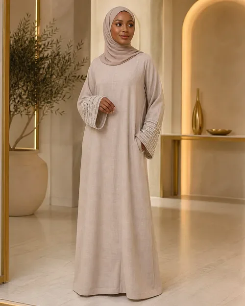 A woman in a beige abaya and hijab standing in a modern, elegant room with decorative vases and a potted plant.
