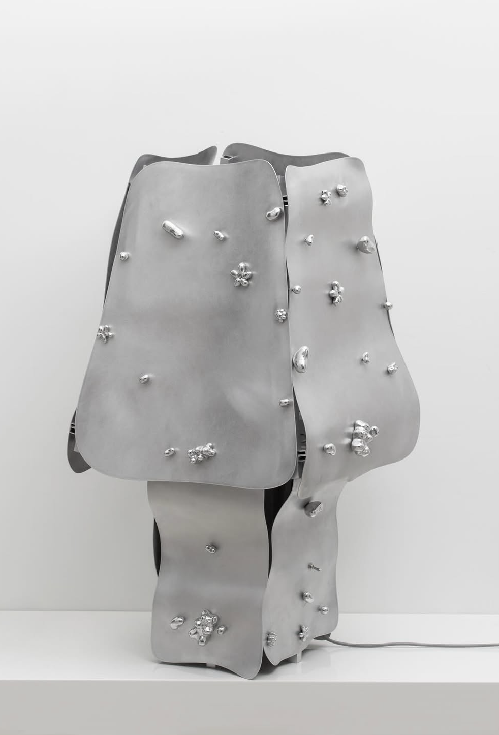 Modern sculpture made of metallic panels with protruding silver nuggets, displayed on a white pedestal against a white background.