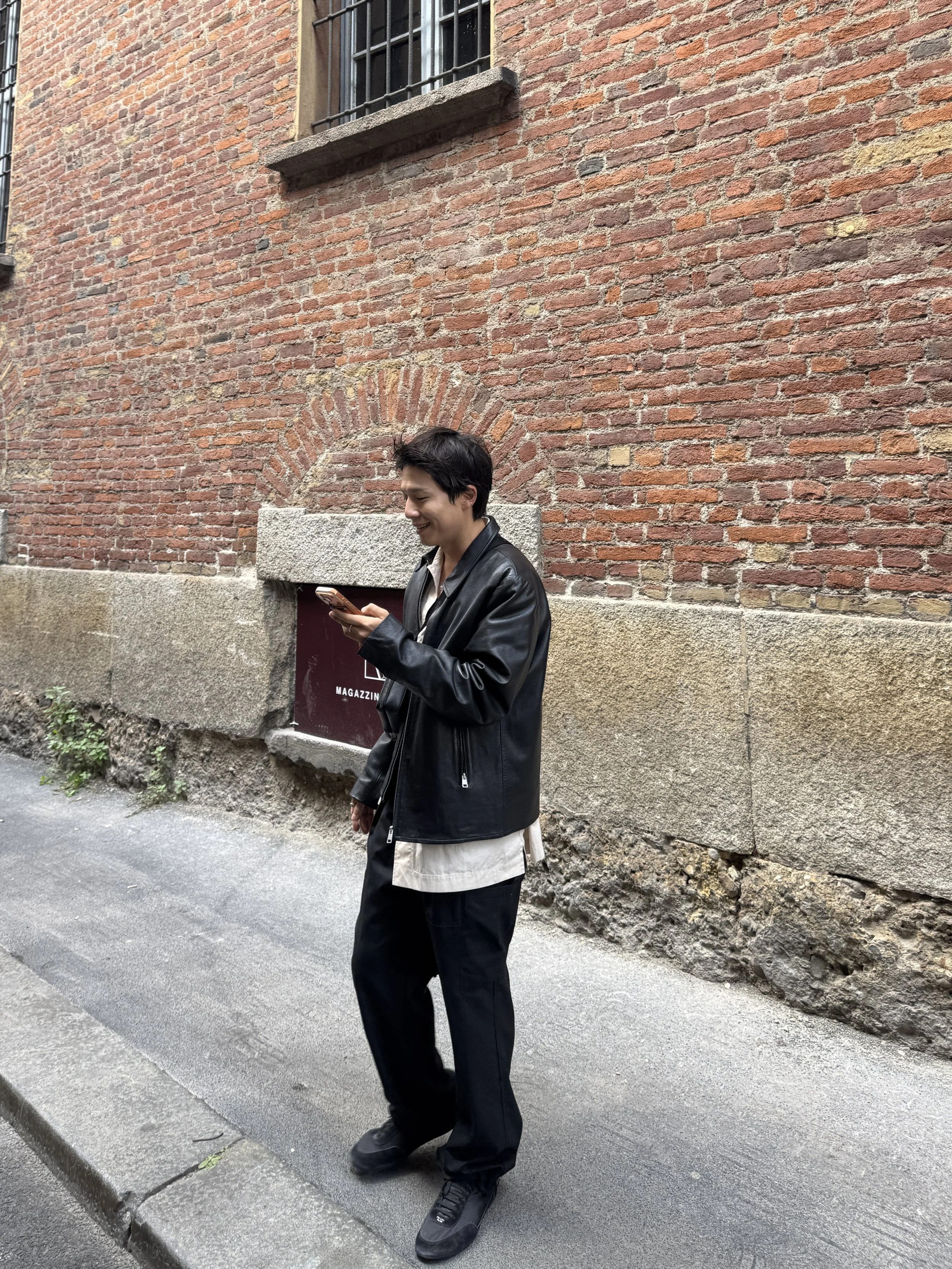 Young man standing on city sidewalk, looking at phone, in black leather jacket, black pants, black sneakers, in front of brick wall with barred window