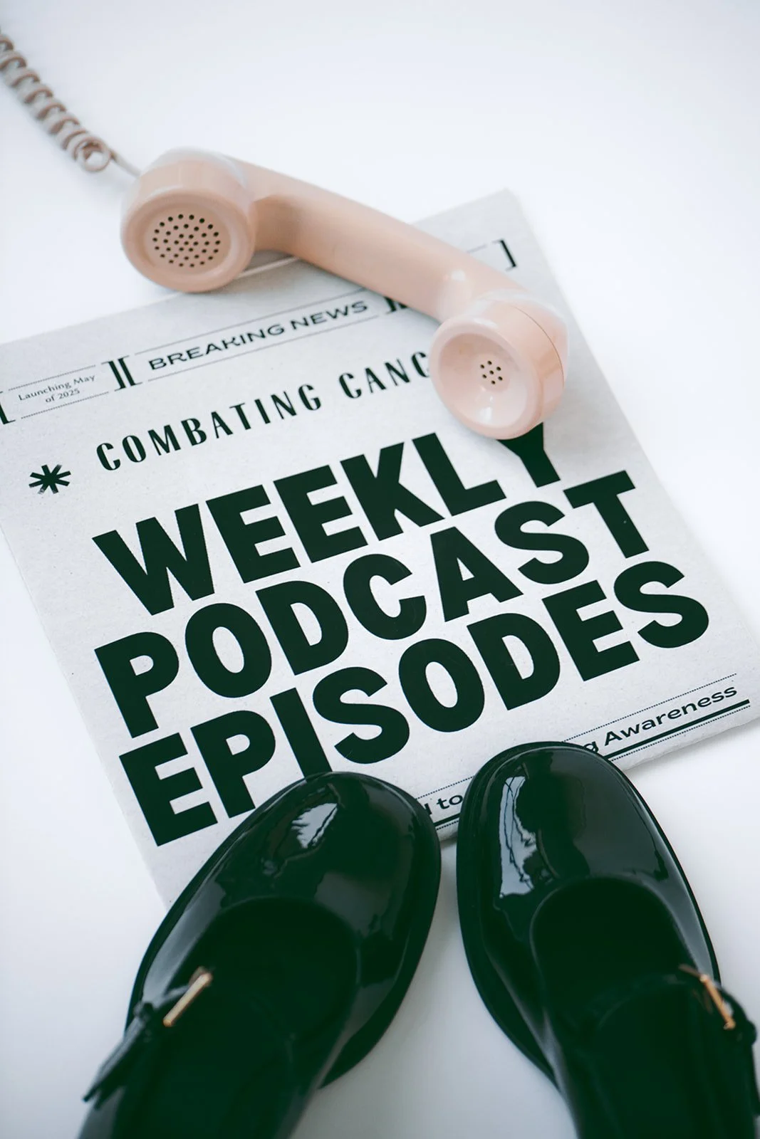 A newspaper with the headline 'Weekly Podcast Episodes' on a white surface, with a pink landline phone placed on top, black shiny shoes, and a pair of white at the top left.