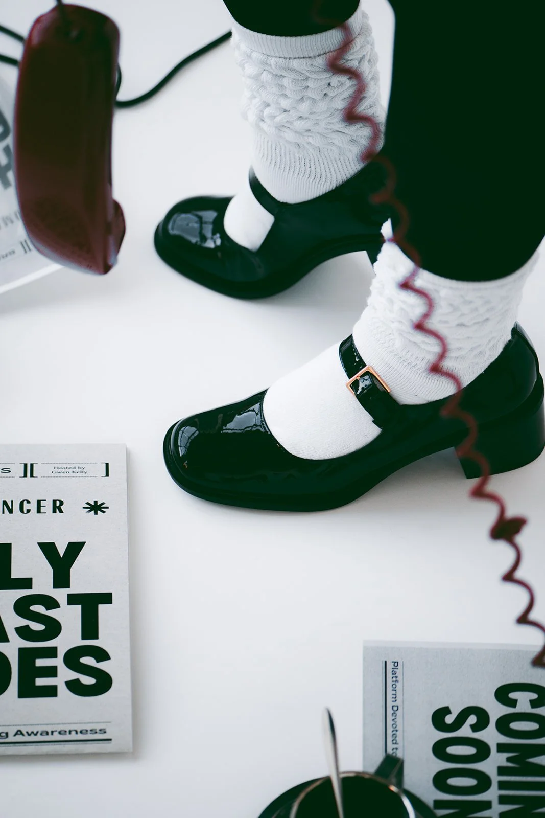A person wearing black patent leather shoes, white socks, white cable-knit leg warmers, and black pants standing on a white surface. Items nearby include a red phone, a black wire, some printed materials, and a cup with a utensil.
