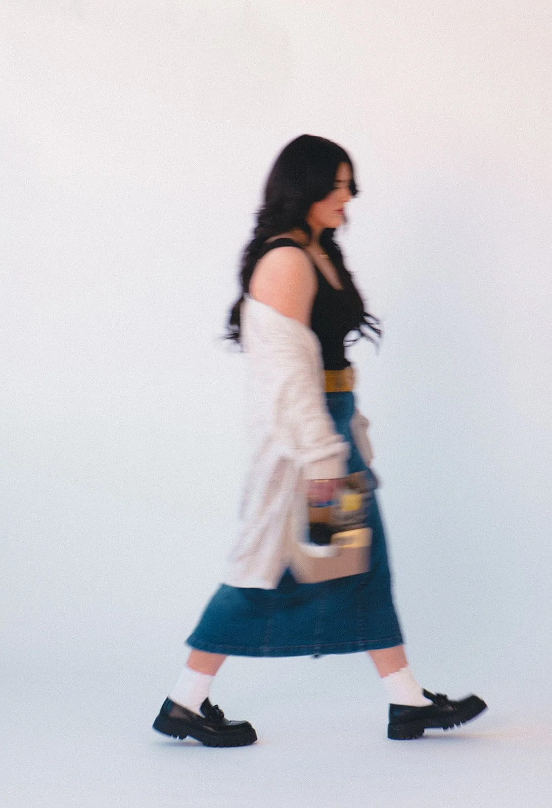 A woman with long dark hair, wearing glasses, a black top, a denim skirt, white socks, and black shoes, walking sideways while holding a beige tote bag with items inside against a plain white background.