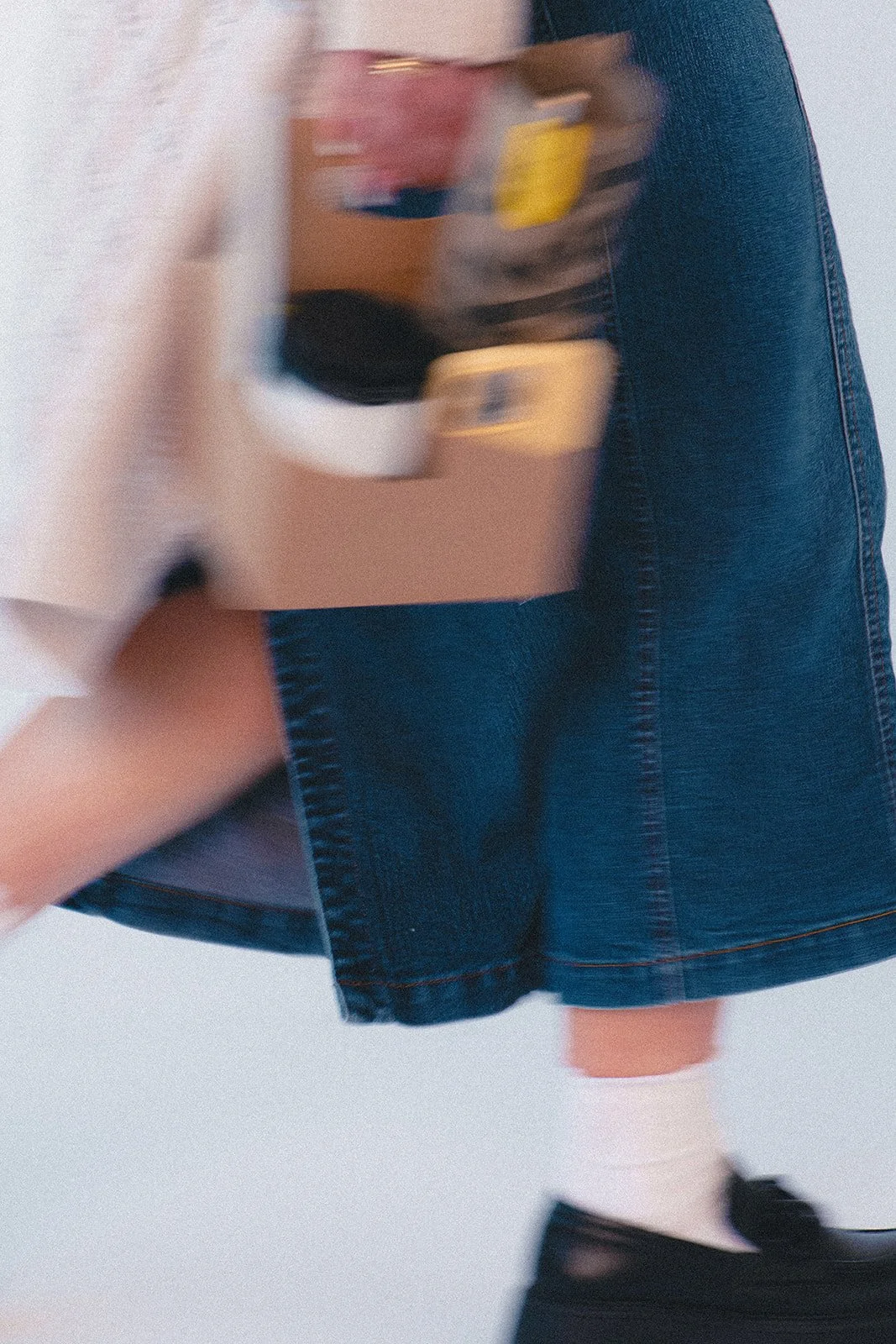 Blurry image of a person walking, wearing a denim skirt, white socks, and black shoes, while holding a colorful object or bag.