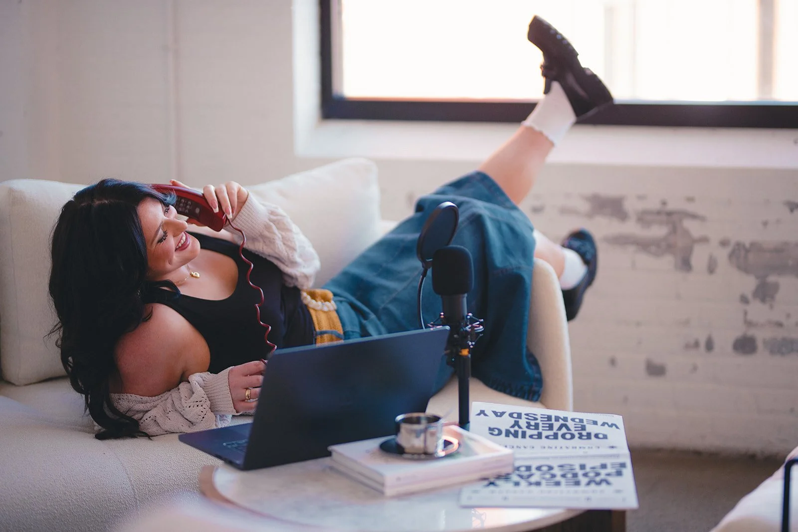 Woman with dark hair lying on a couch speaking on a corded phone, smiling, with one leg resting on the windowsill. She is wearing a black top, a cream-colored cardigan, and denim shorts. A laptop and a book titled 'Dropping Every Day' are on the table in front of her.
