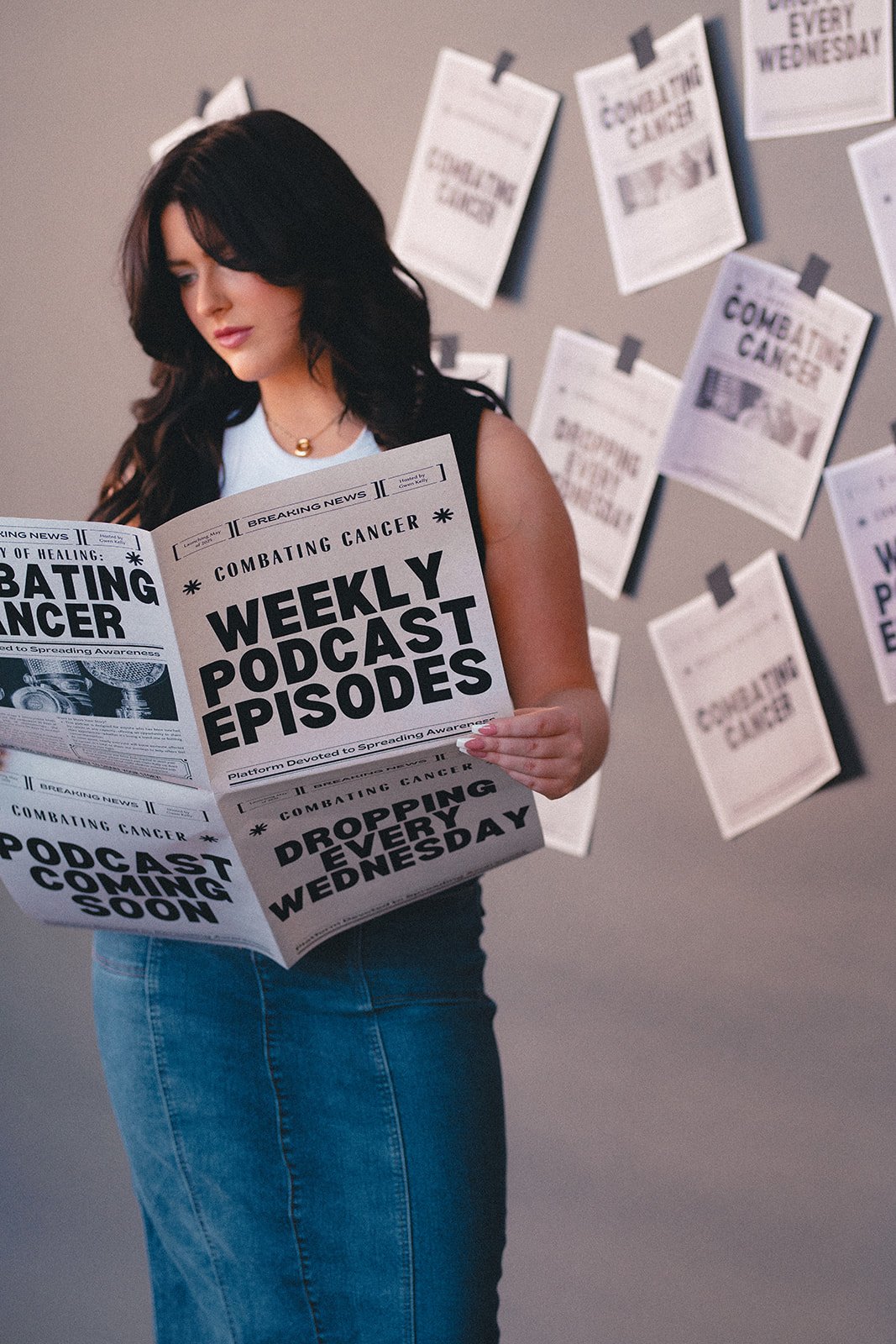 A woman with dark hair and a white sleeveless top reading a newspaper with headlines about cancer awareness and podcasts, standing in front of a wall decorated with similar newspapers.