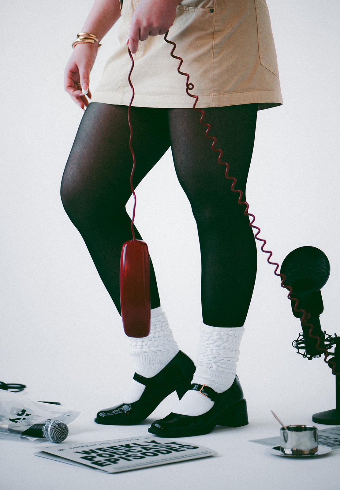 Close-up of a person standing with one foot forward, wearing black tights, white socks, black shoes, and a beige skirt, holding a vintage red corded telephone. Items on the ground include a microphone, newspapers, a teacup with a saucer, and a vintage microphone on a stand.