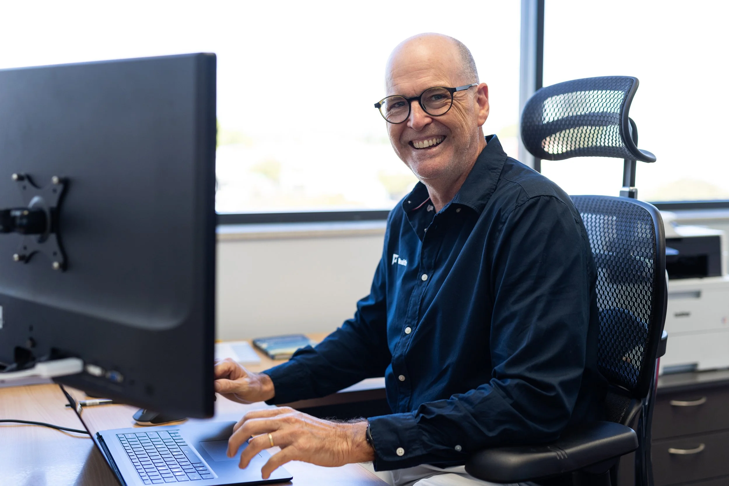 Smiling man with glasses working at a desk in an office using a computer.