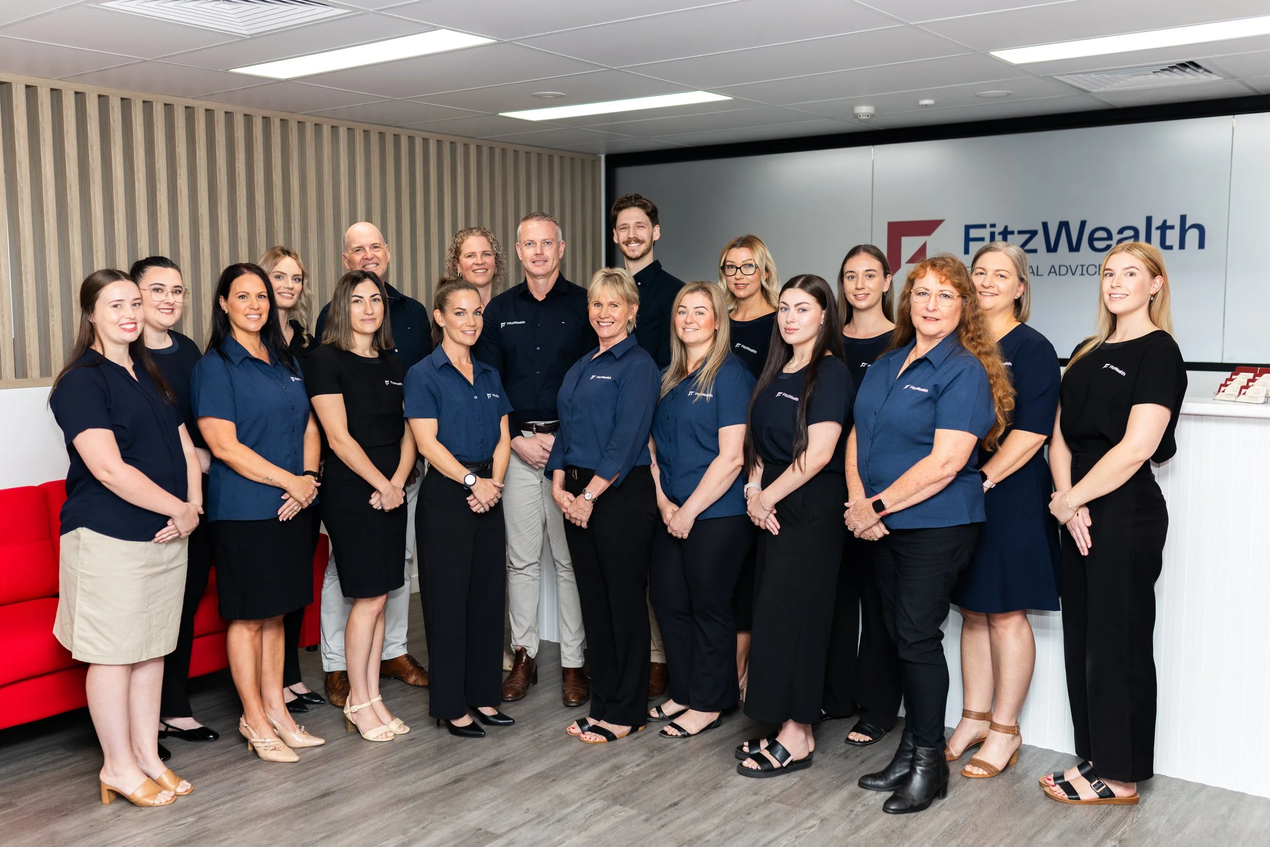 Group of professional people posing for a photo in an office setting, standing in front of a sign that reads 'FitzWealth'.