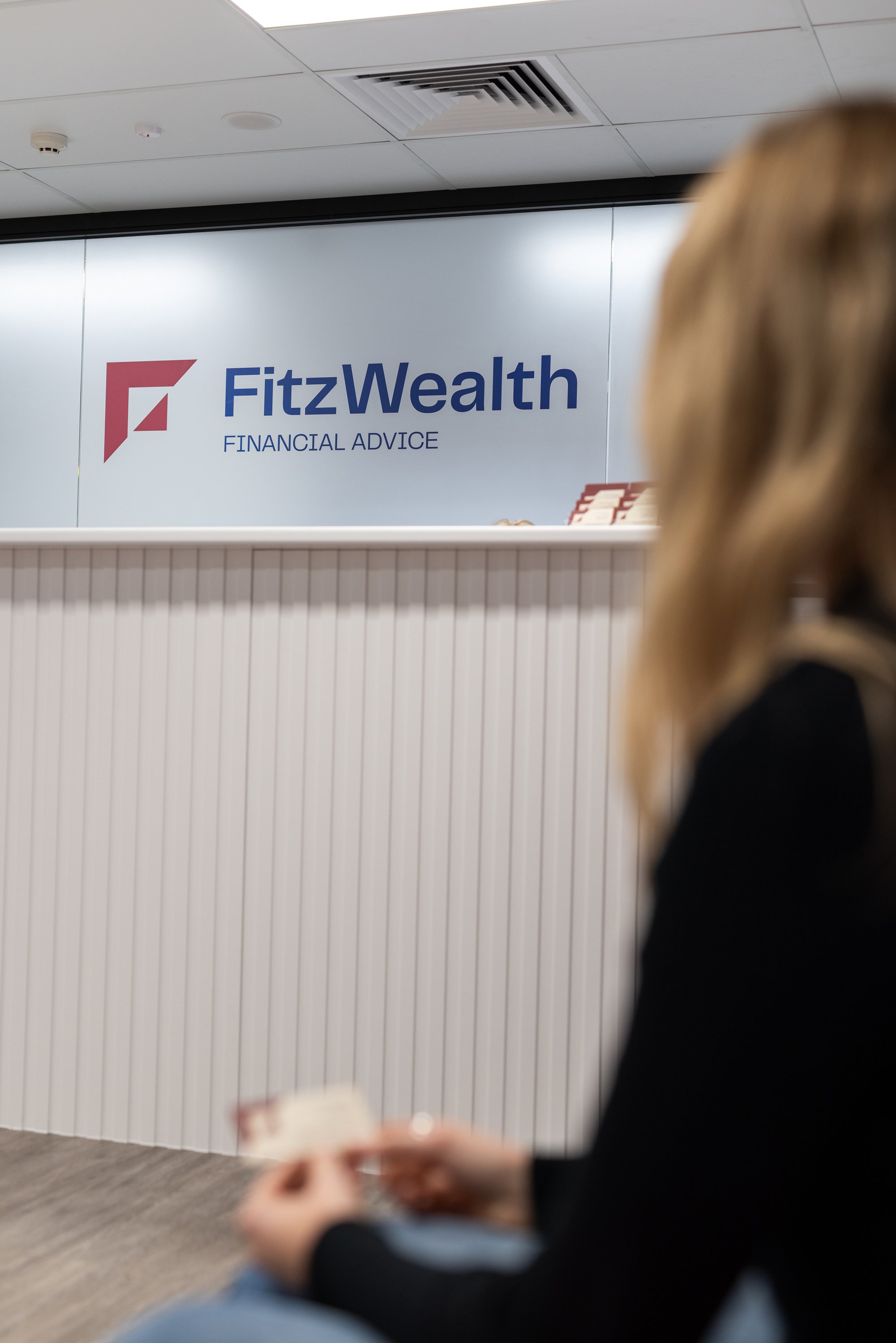 A woman sitting in front of a FitzWealth Financial Advice sign at an indoor office or reception area.