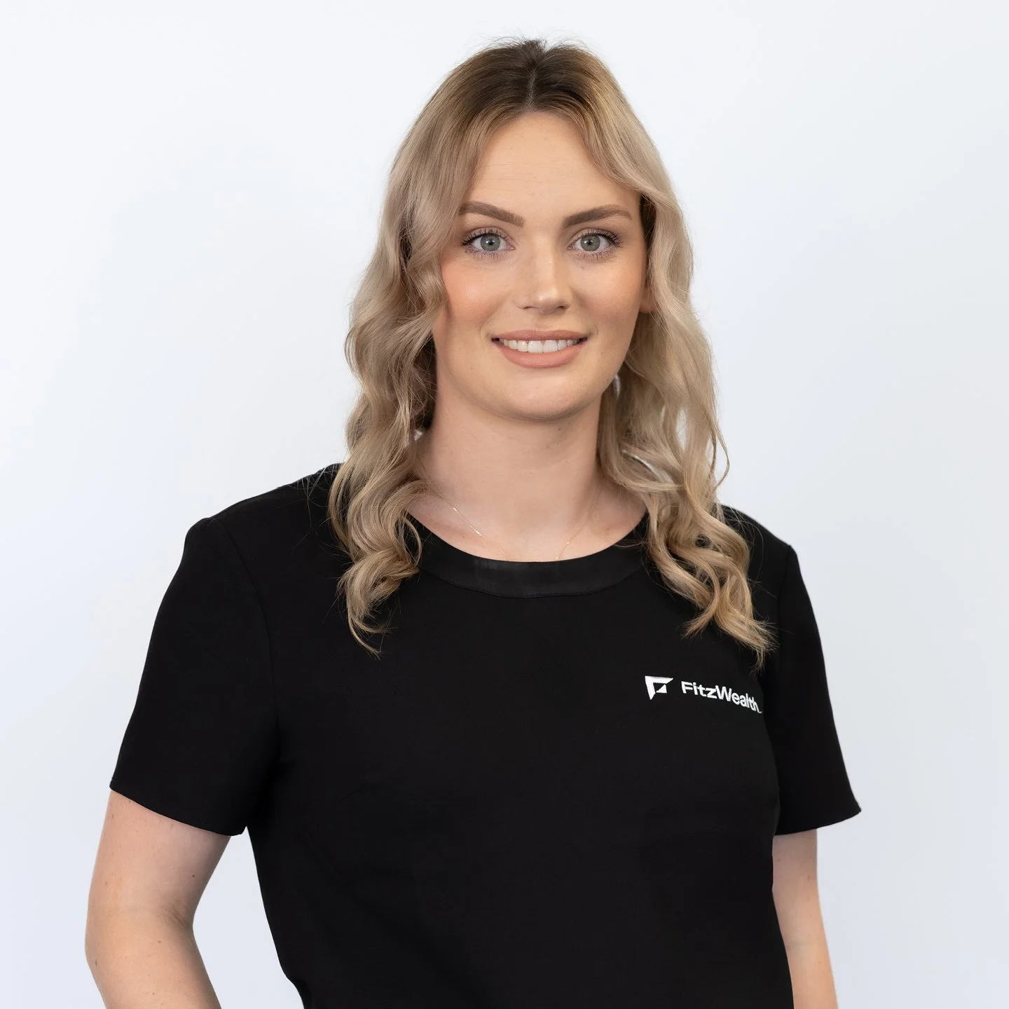 A woman with blonde, wavy hair, blue eyes, and fair skin, wearing a black FitzWellth T-shirt, smiling against a plain white background.
