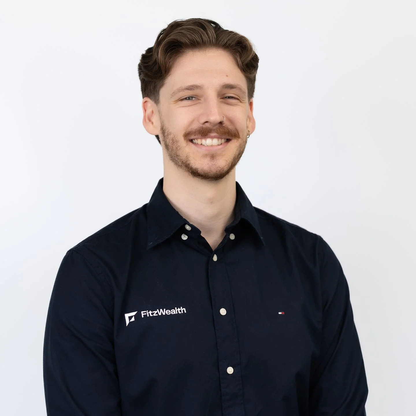 A young man with light brown hair and facial hair, smiling, wearing a dark blue button-up shirt with the Fitz Wealth logo, standing against a plain light background.