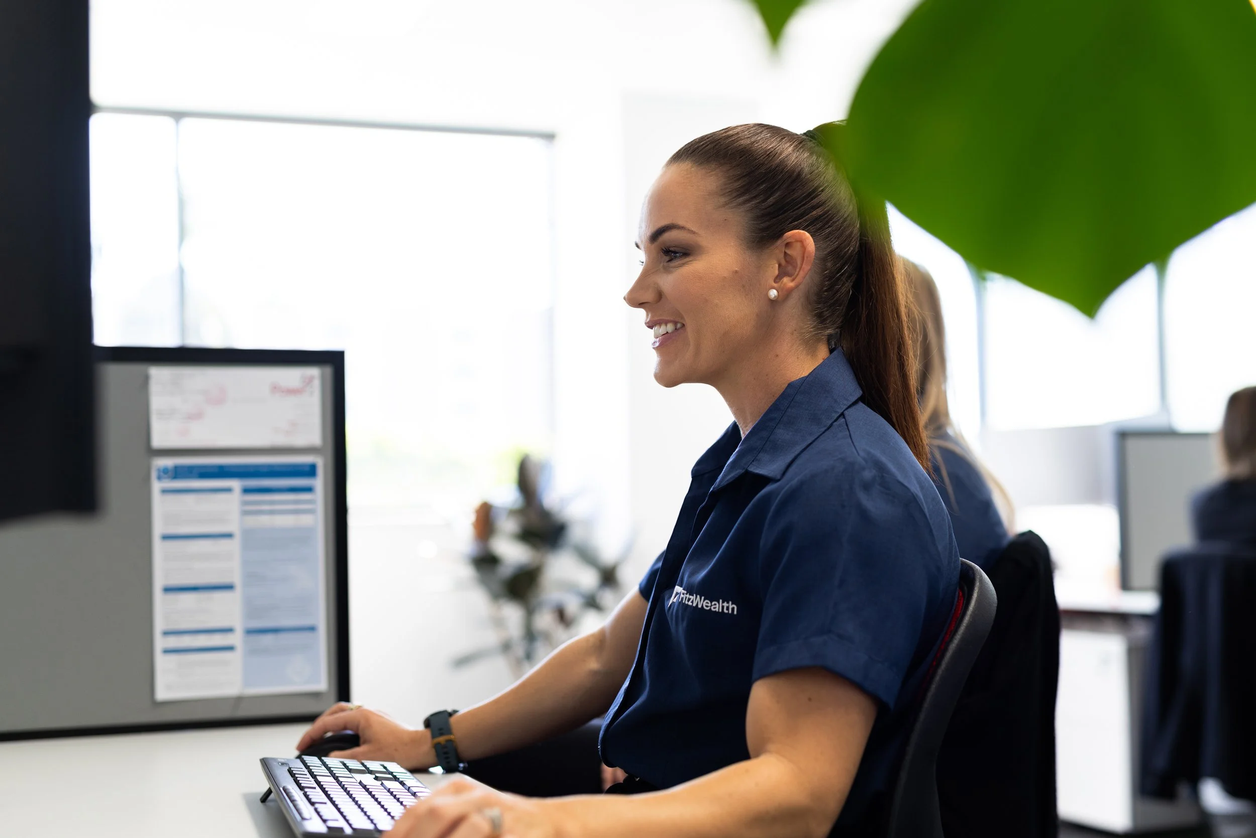 A woman smiling while working at a computer in an office, wearing a blue shirt with 'Fitz Wealth' embroidered on it.