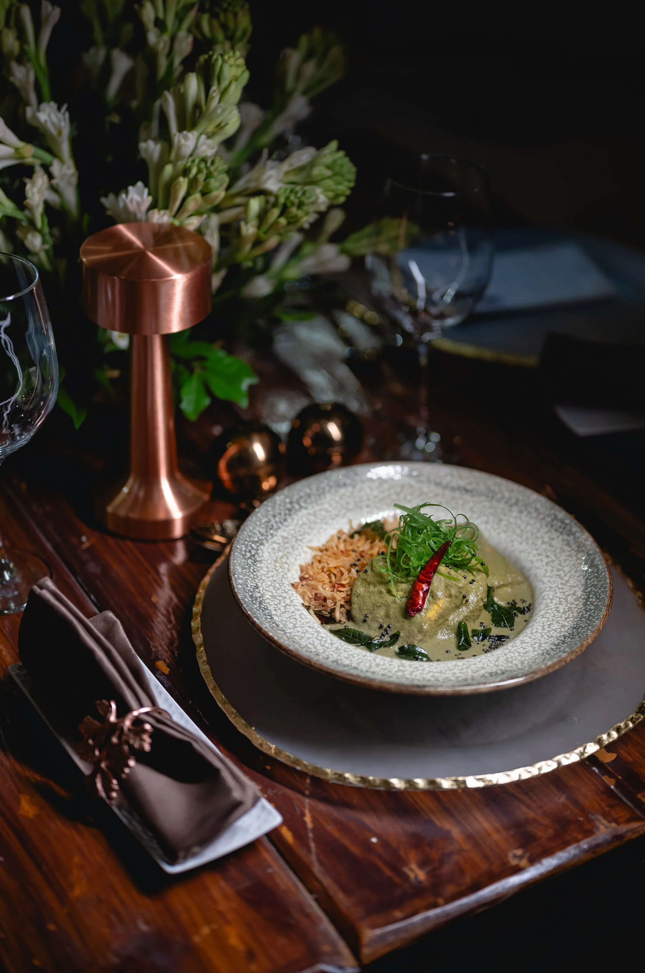 A gourmet dish on a decorative plate with rice, a green sauce, chili pepper, and garnishes, set on a dark wooden table with wine glasses, a floral centerpiece, and elegant tableware.