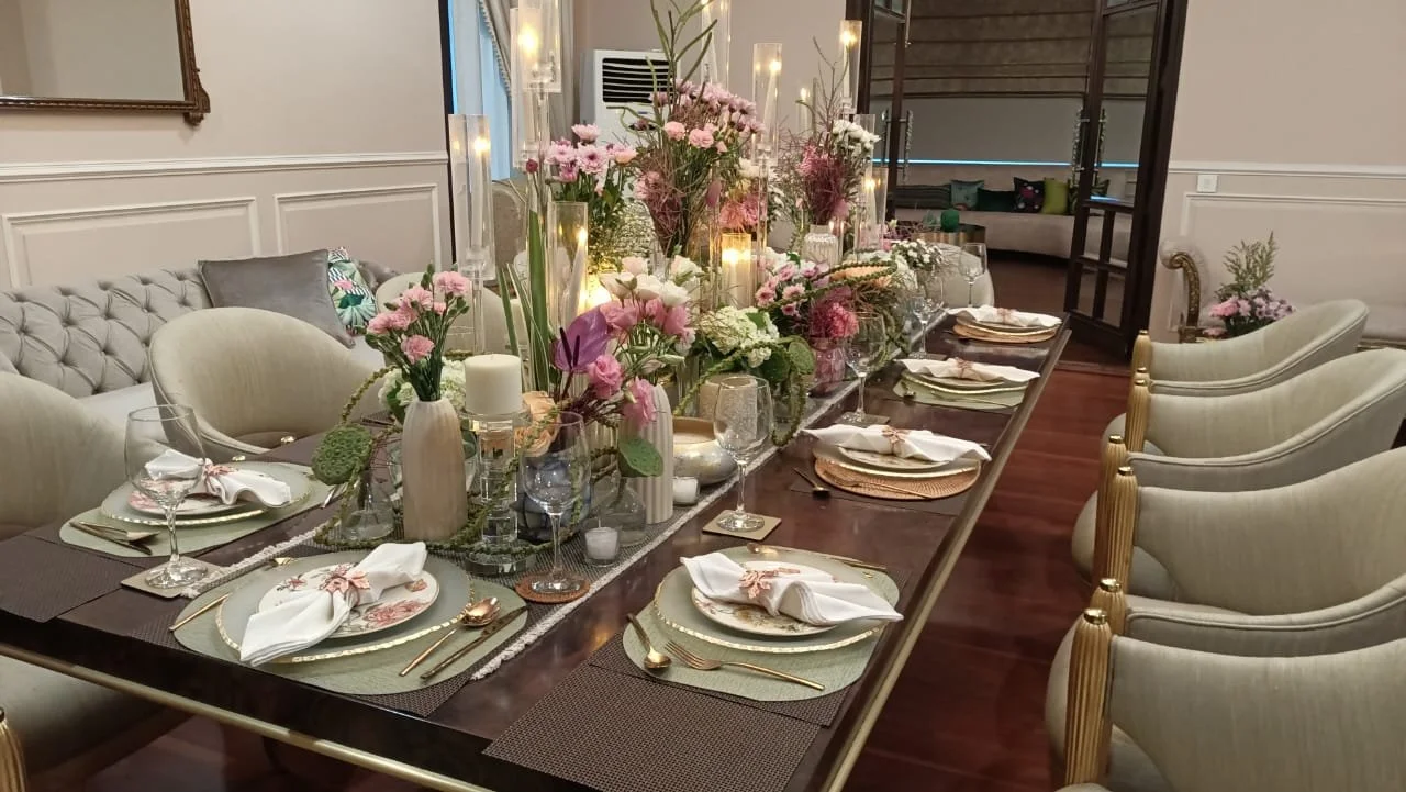 A dining table set for a meal with floral centerpieces, candles, plates, cloth napkins, and glassware in a decorated room.