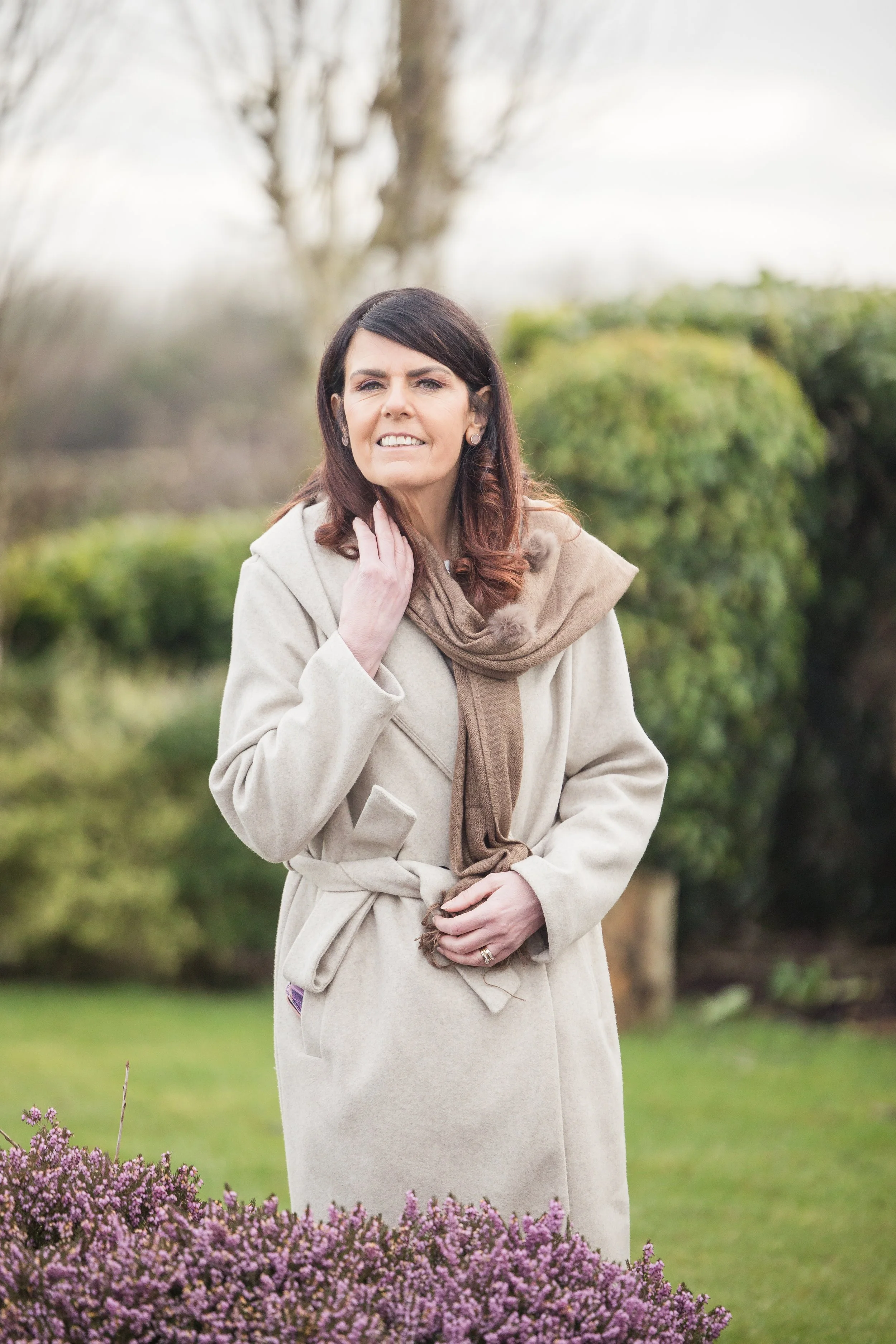 Woman outdoors wearing a beige coat and scarf, with greenery in the background.