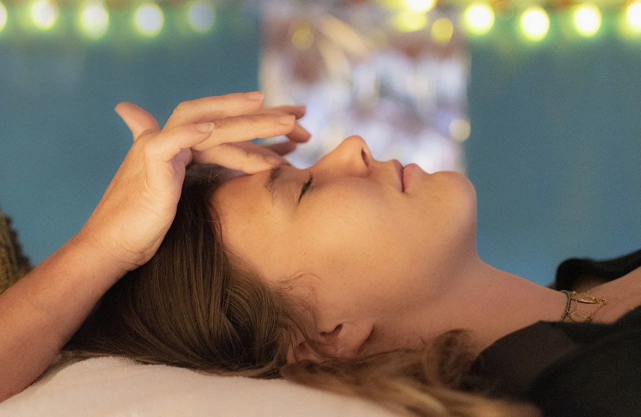 Person receiving a relaxing forehead massage, lying down with closed eyes, blurred lights in the background.
