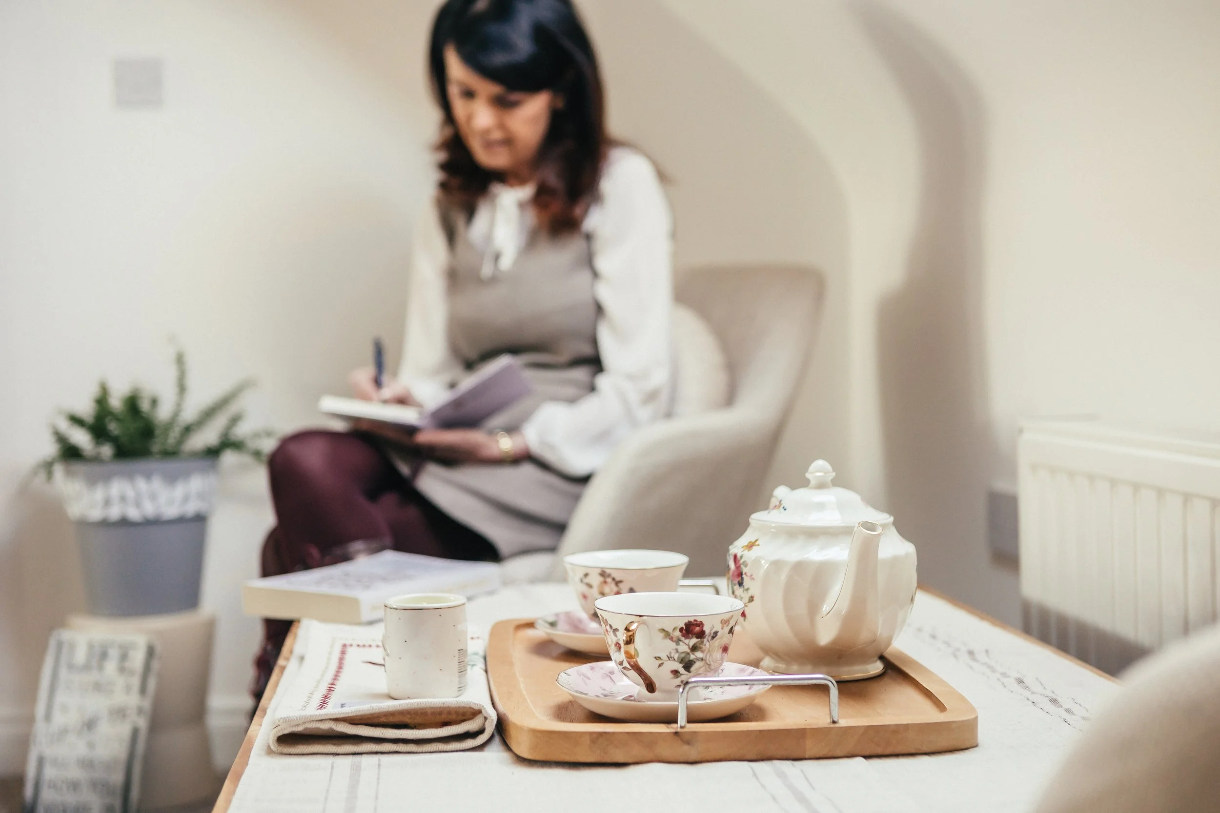 A woman writing in a notebook while sitting in an armchair, with a wooden tray holding a floral teapot and teacups on a table in the foreground.