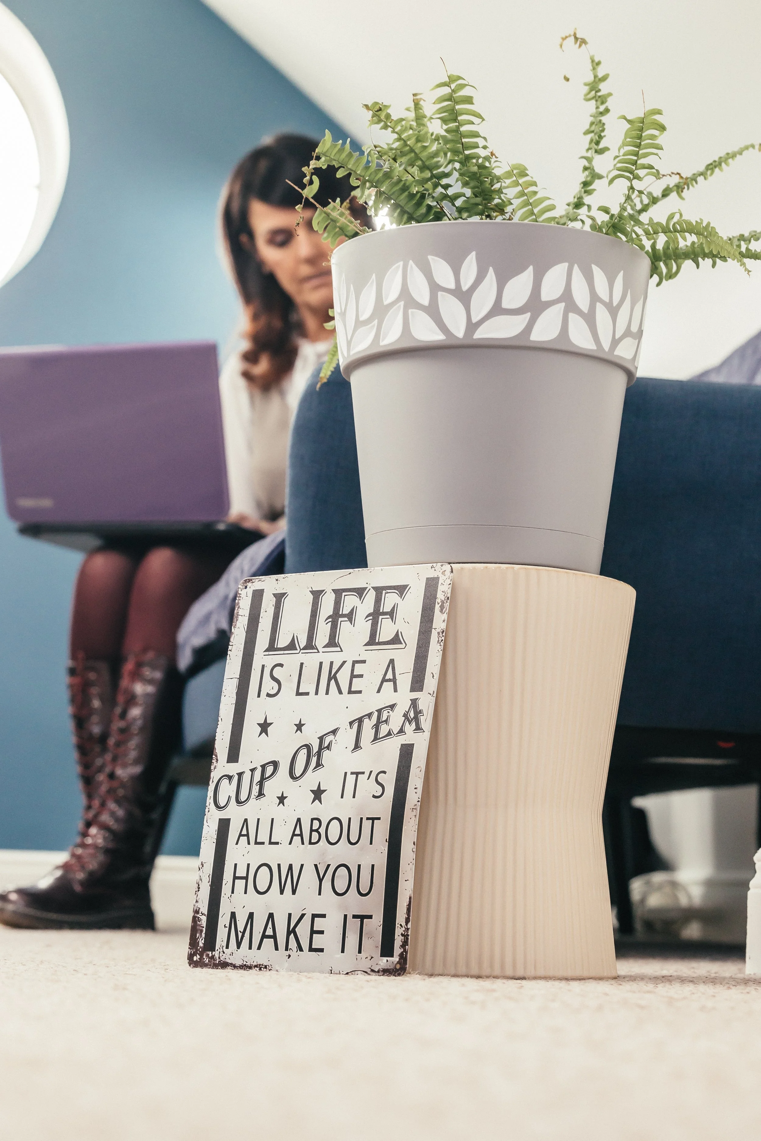 A person sitting on a couch with a laptop and a sign leaning on a stand. The sign reads, 'Life is like a cup of tea. It's all about how you make it.' There is a fern plant in a decorative pot next to the sign.