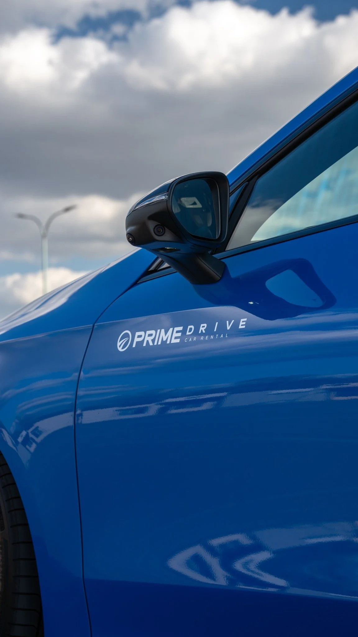Close-up of a blue car with Prime Drive Car Rental logo, focusing on the side mirror and door, with a cloudy sky background.