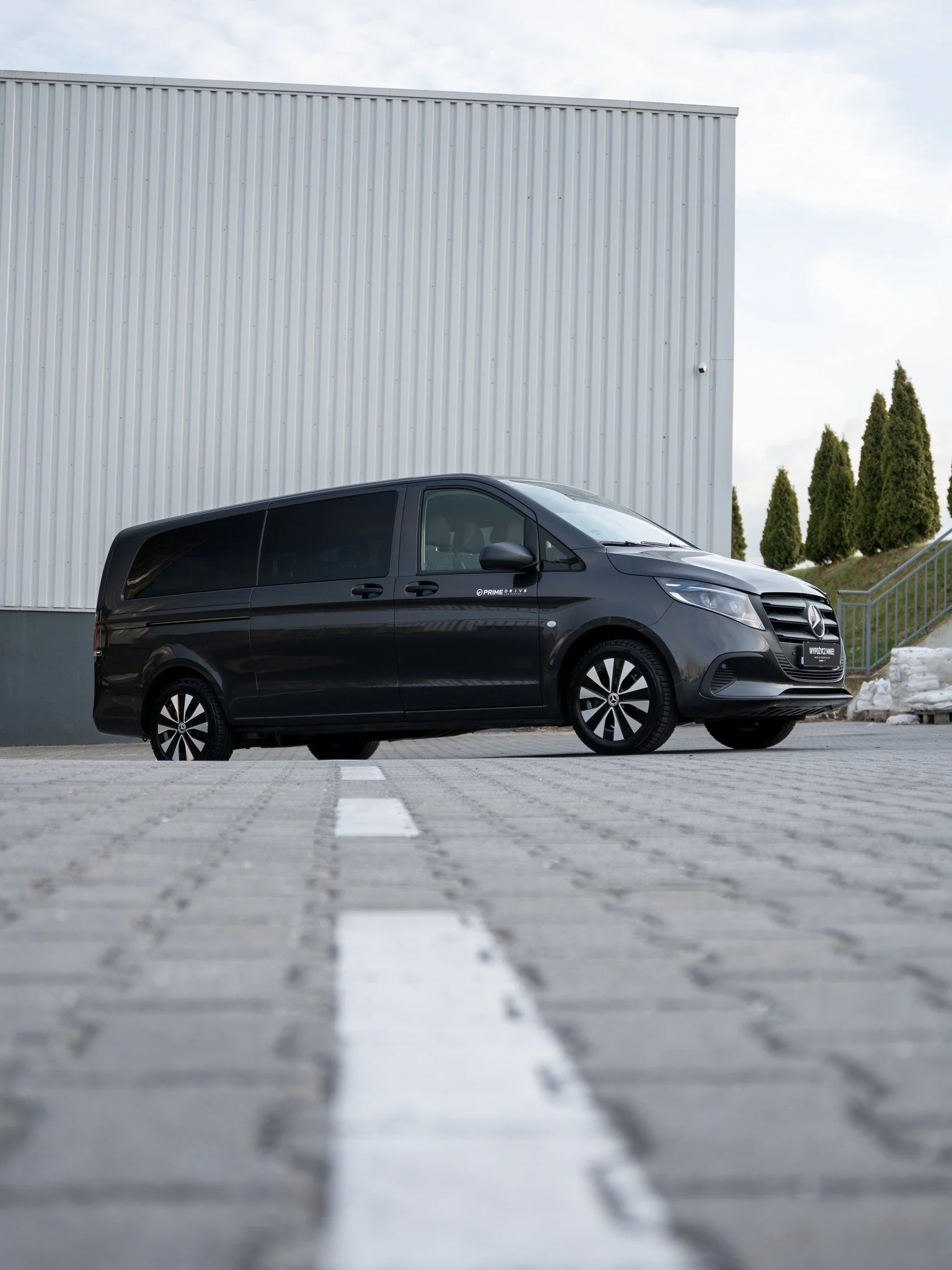 Black Mercedes-Benz van parked in an empty parking lot with a large industrial building and trees in the background.