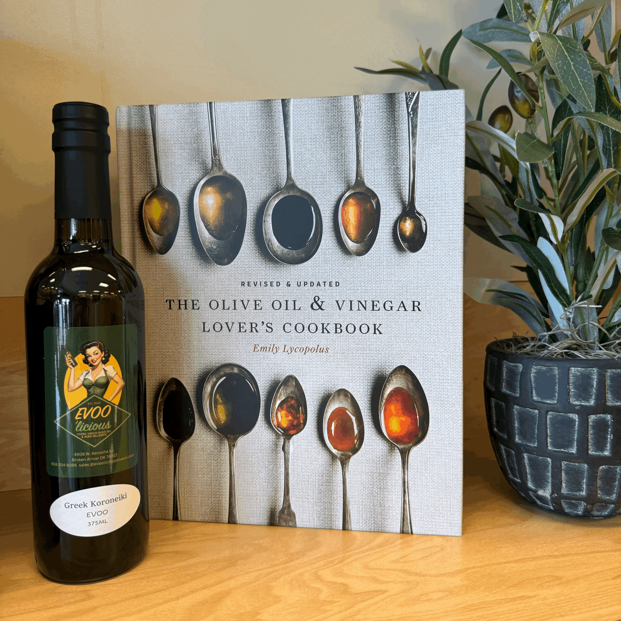 A close-up photo of a cookbook titled "The Olive Oil & Vinegar Lover's Cookbook" by Emily Lycopolus, featuring images of various spoons with oil and vinegar samples, with a bottle of olive oil with a vintage pin-up style woman on the label and a potted plant in the background.