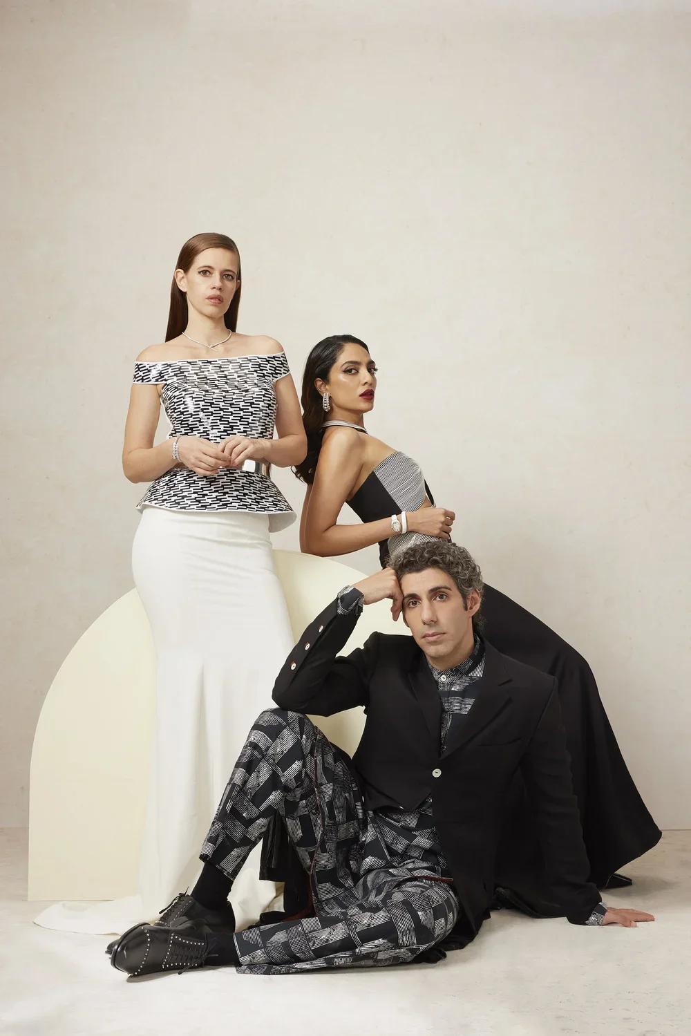 Three fashionably dressed individuals, two women and one man, posed against a plain background with minimal decor. The woman on the left is standing with her hands clasped in front of her, wearing a patterned off-the-shoulder top and a long white ski