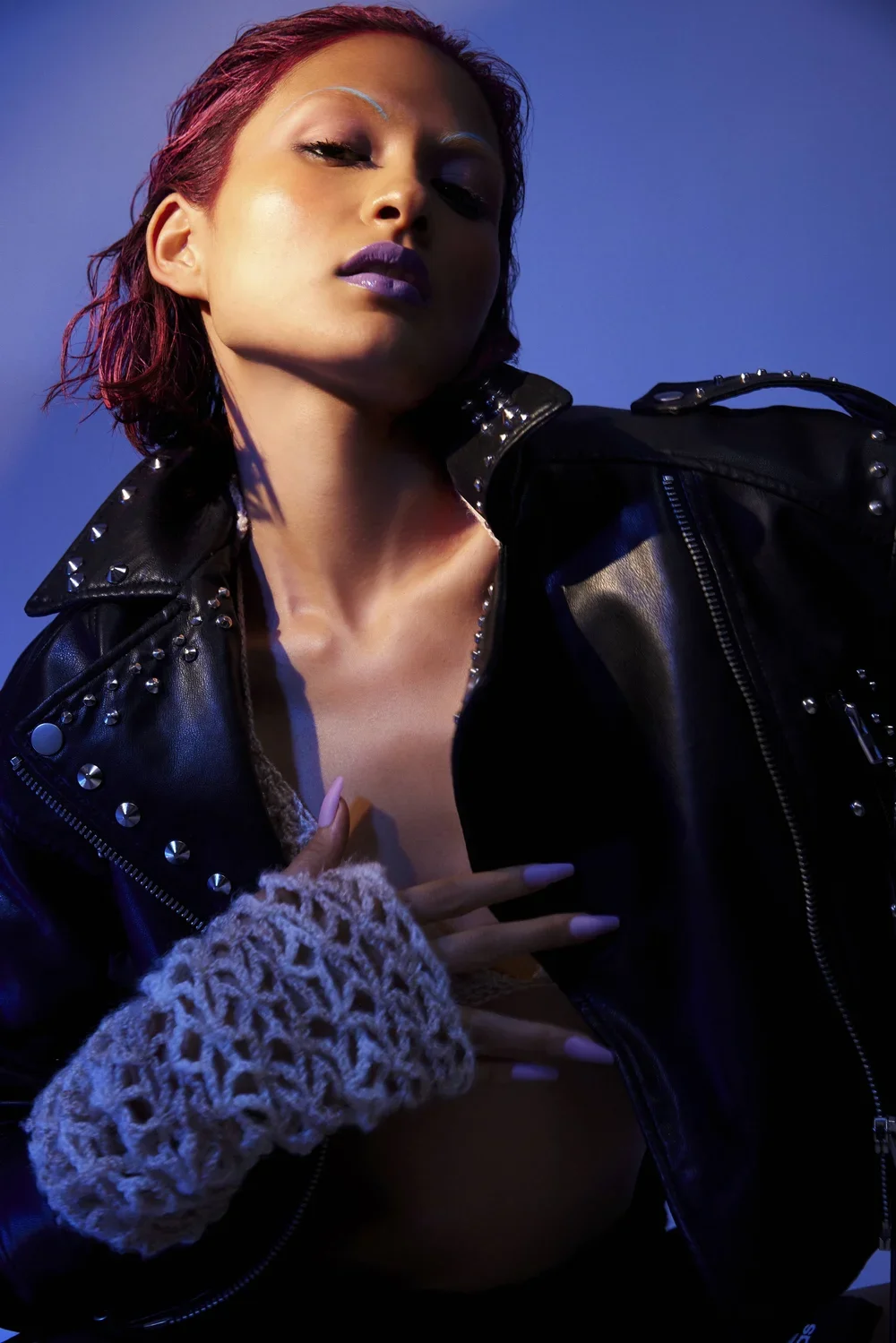A woman with short red hair and purple lipstick, wearing a black leather jacket with metal studs, posing with her eyes closed against a blue background.