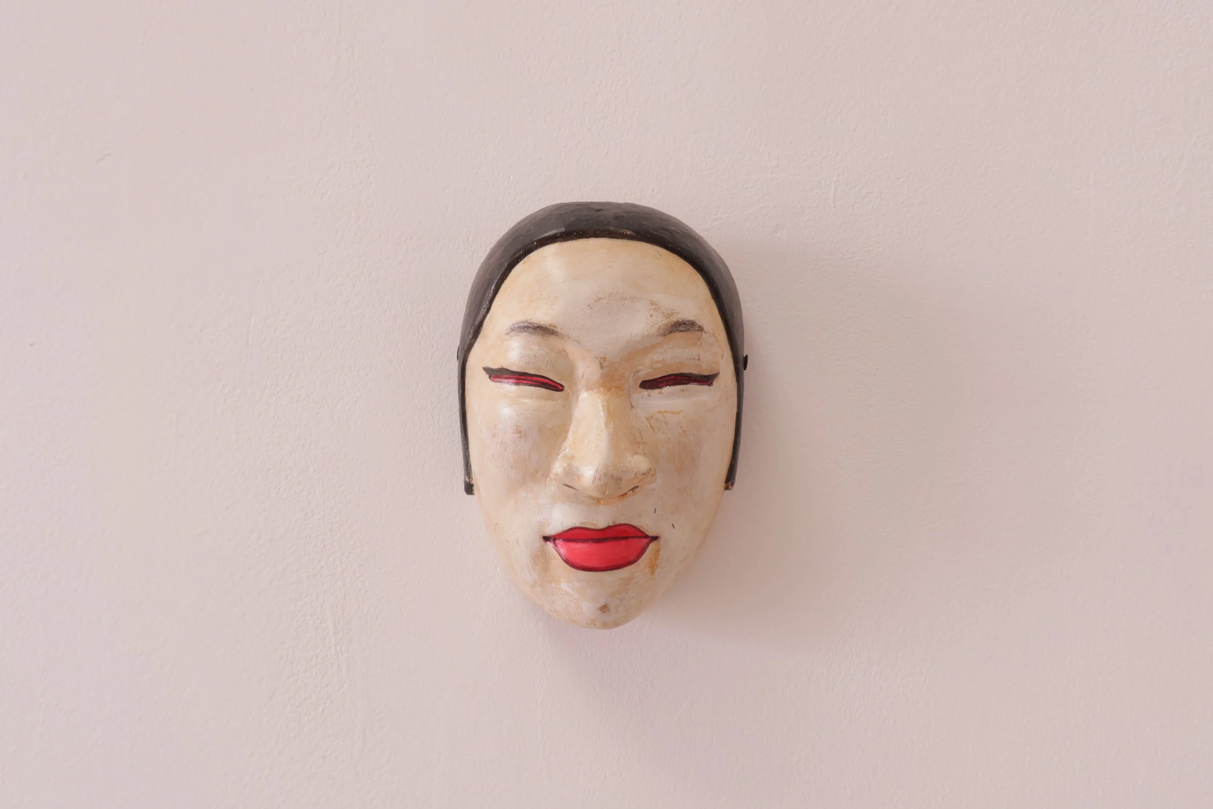 Decorative mask of a smiling woman with closed eyes, red lips, and black hair against a plain wall.