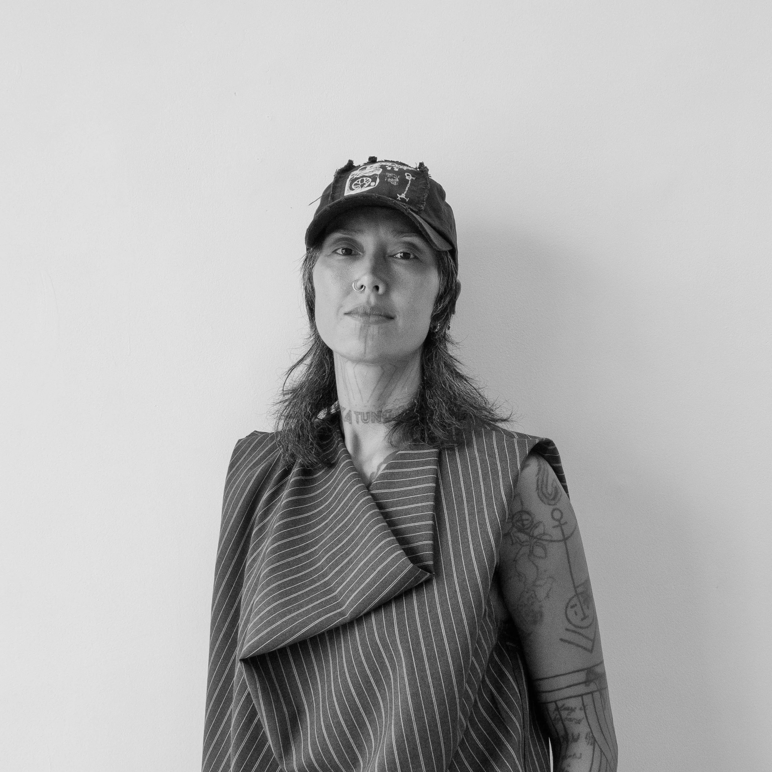 Black and white photo of a woman with tattoos, wearing a baseball cap and striped sleeveless top, standing against a plain wall.