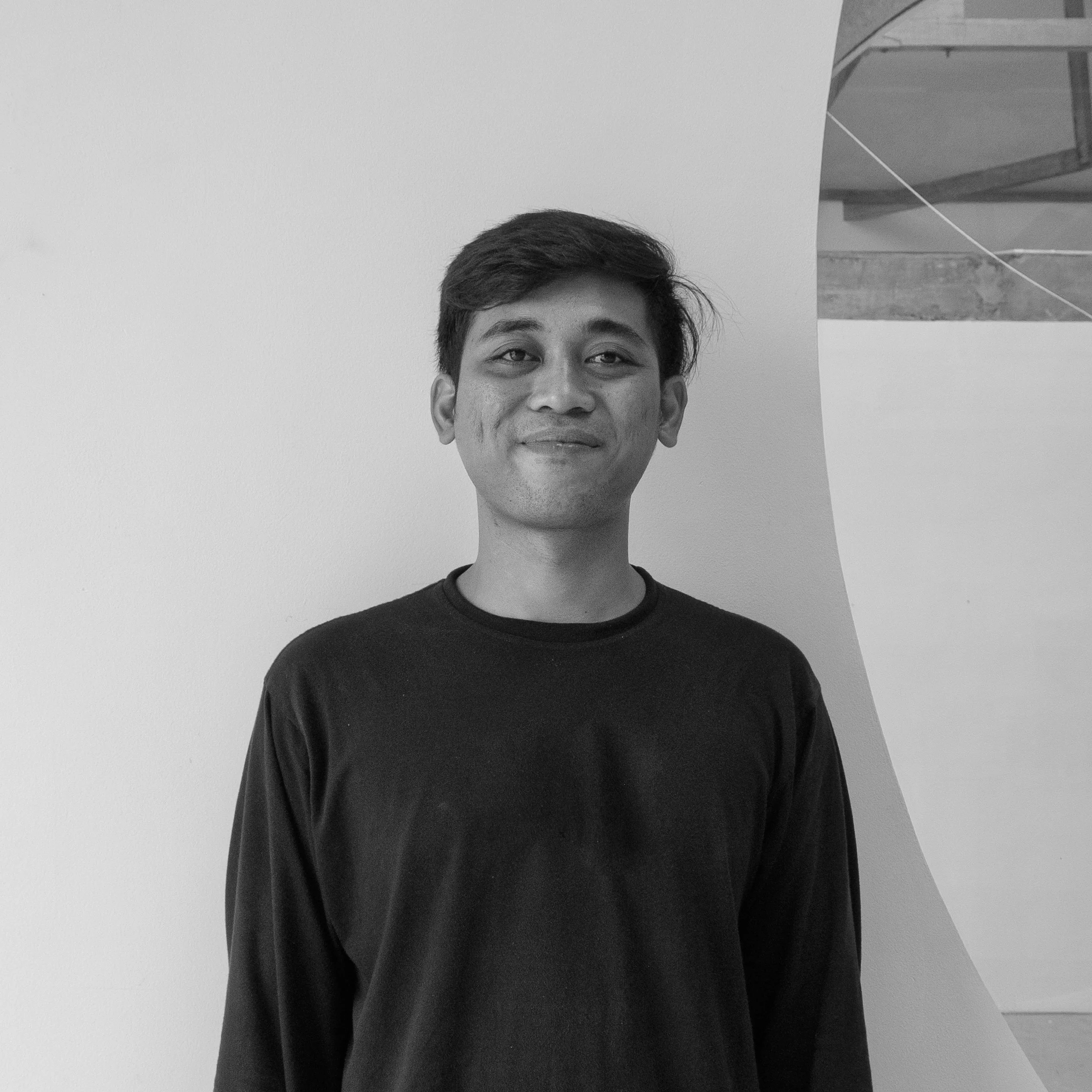 Black and white photo of a young man with short hair, smiling slightly, wearing a black long sleeve shirt, standing in front of a plain wall with a curved architectural feature in the background.