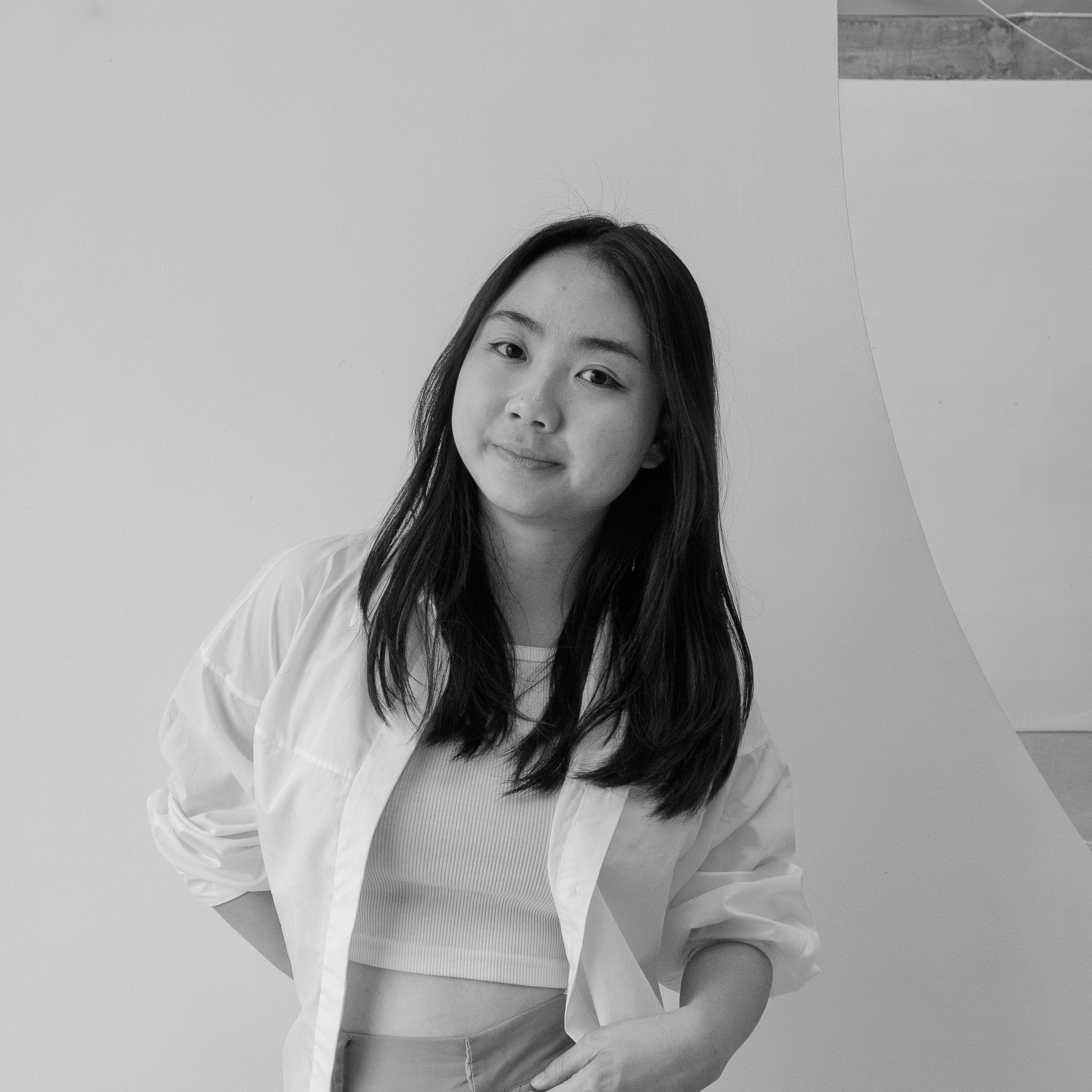 A young woman with long dark hair, wearing a white shirt over a light-colored top, standing in front of a plain wall with some geometric shapes in the background.