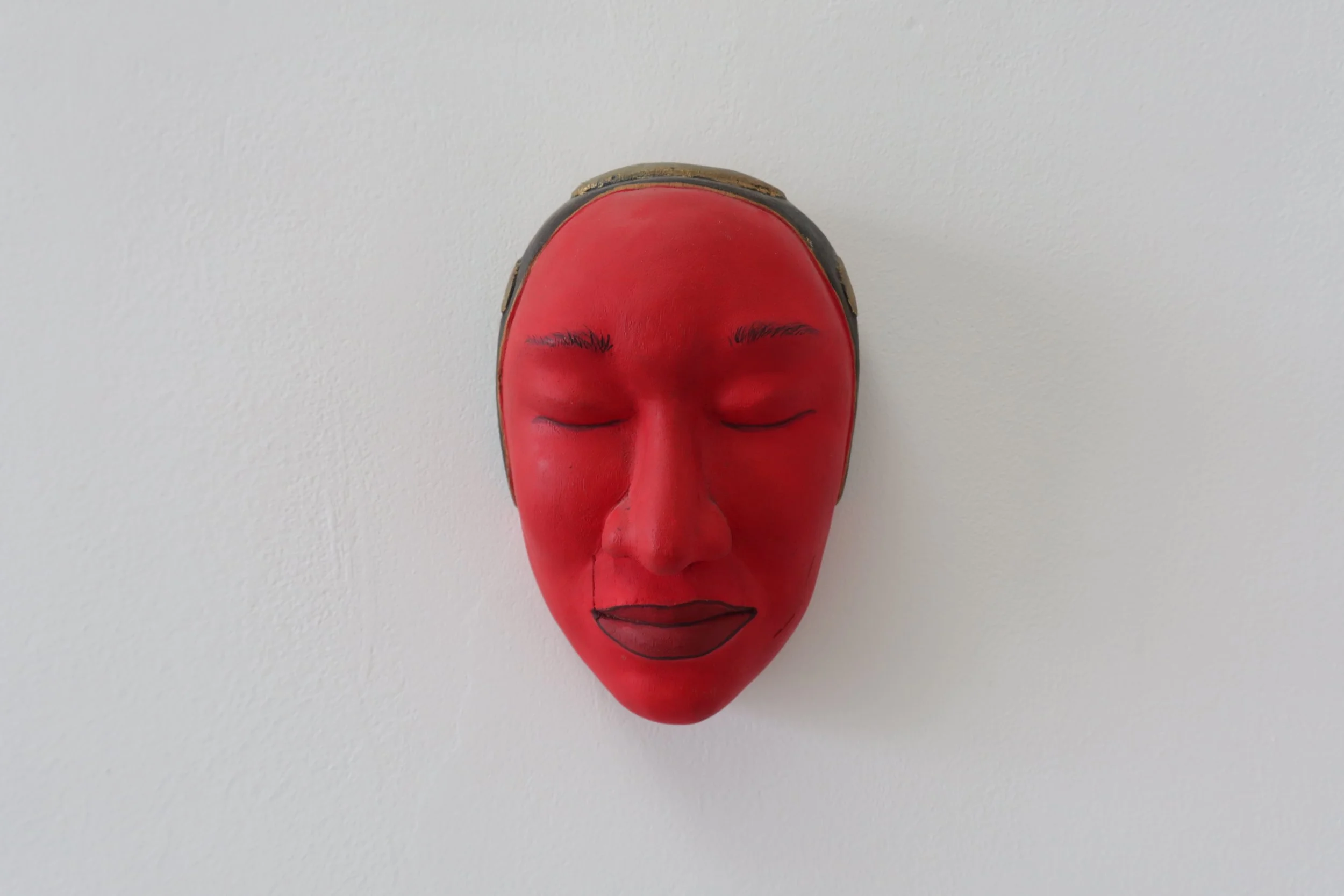 A red mask of a woman's face with closed eyes and black eyebrows, mounted on a white wall.