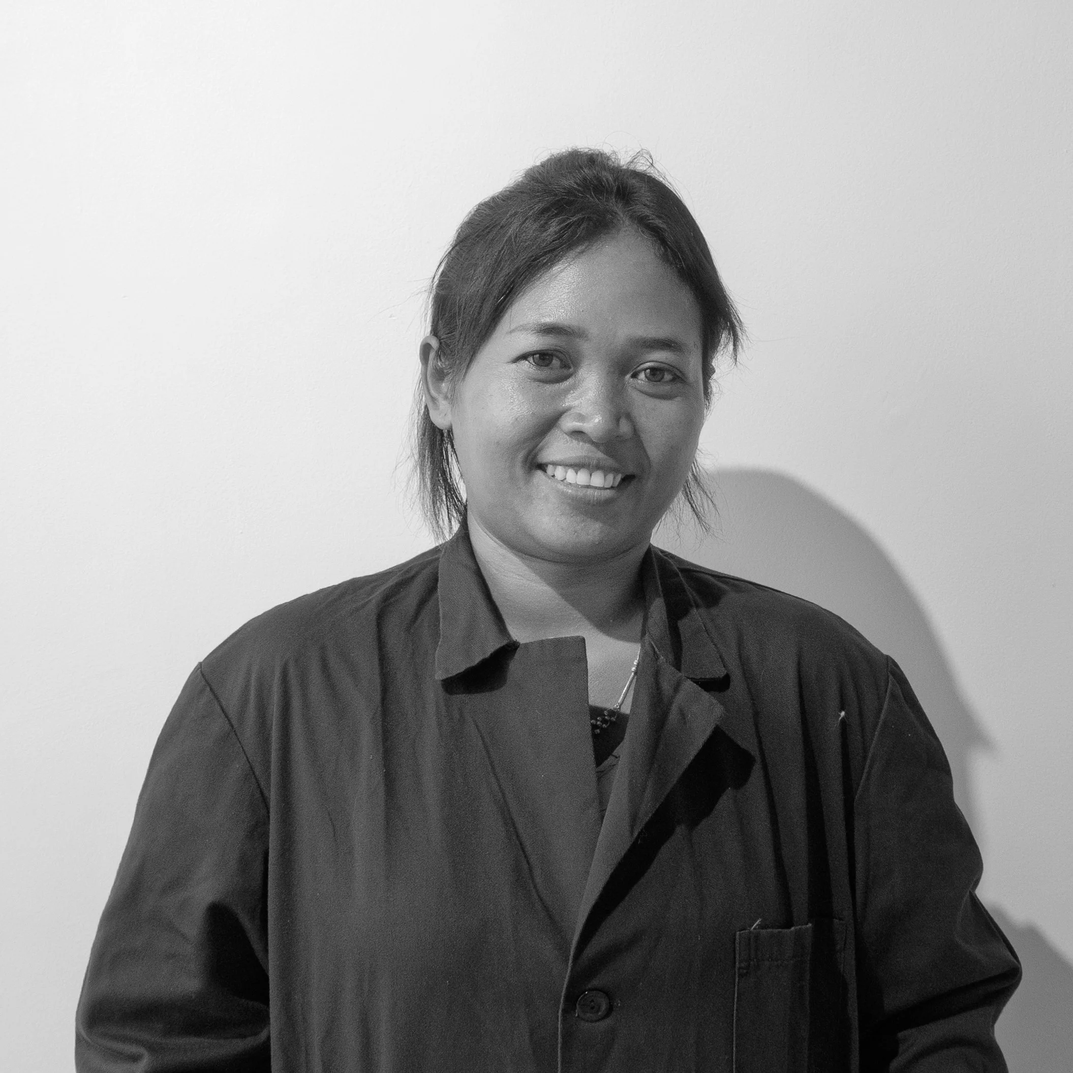 Black and white portrait of a woman with short hair, smiling, wearing a dark shirt with a collar, standing in front of a plain light-colored wall.