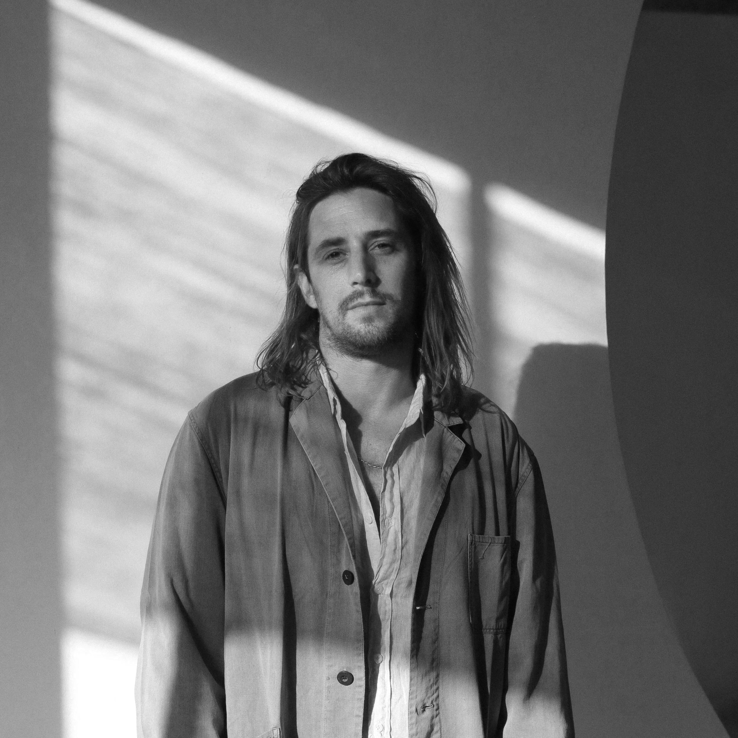 Male with shoulder-length hair and facial hair standing in front of a wall with shadows and geometric shapes.